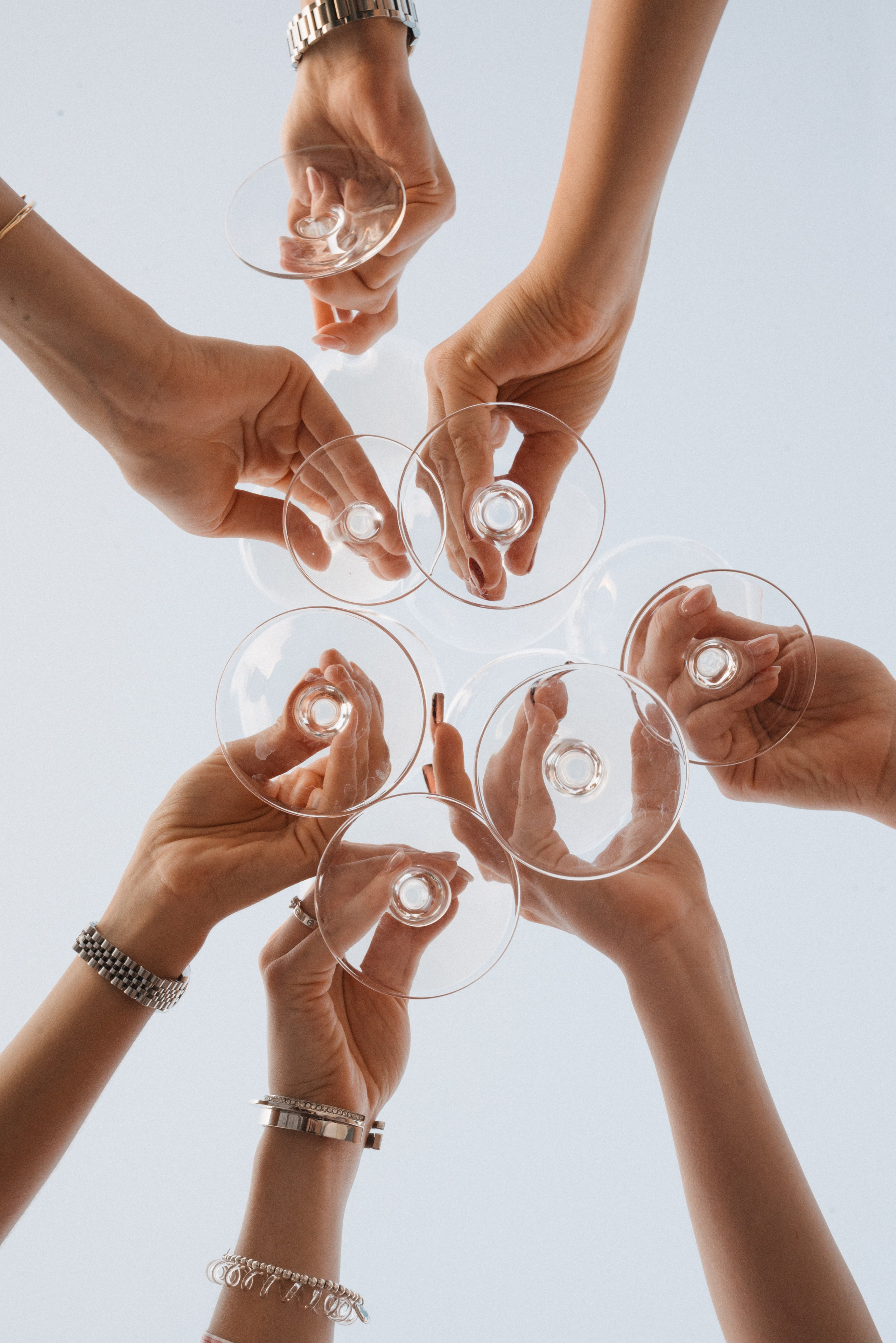 Снимок семи рук девушек снизу в процессе чоканья прозрачными бокалами на фоне неба