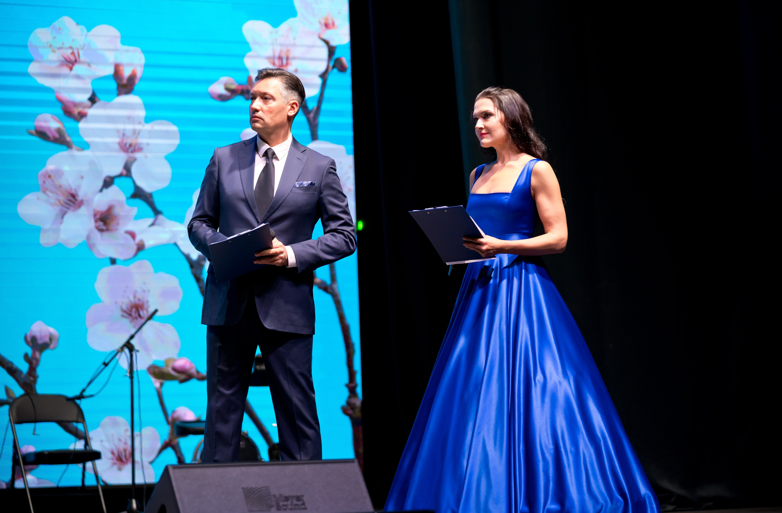 Ведущие мероприятия: мужчина в тёмно-синем костюме и женщина в ярком синем платье на сцене. День местного самоуправления, Санкт-Петербург. Фото Евгения Мальцева.