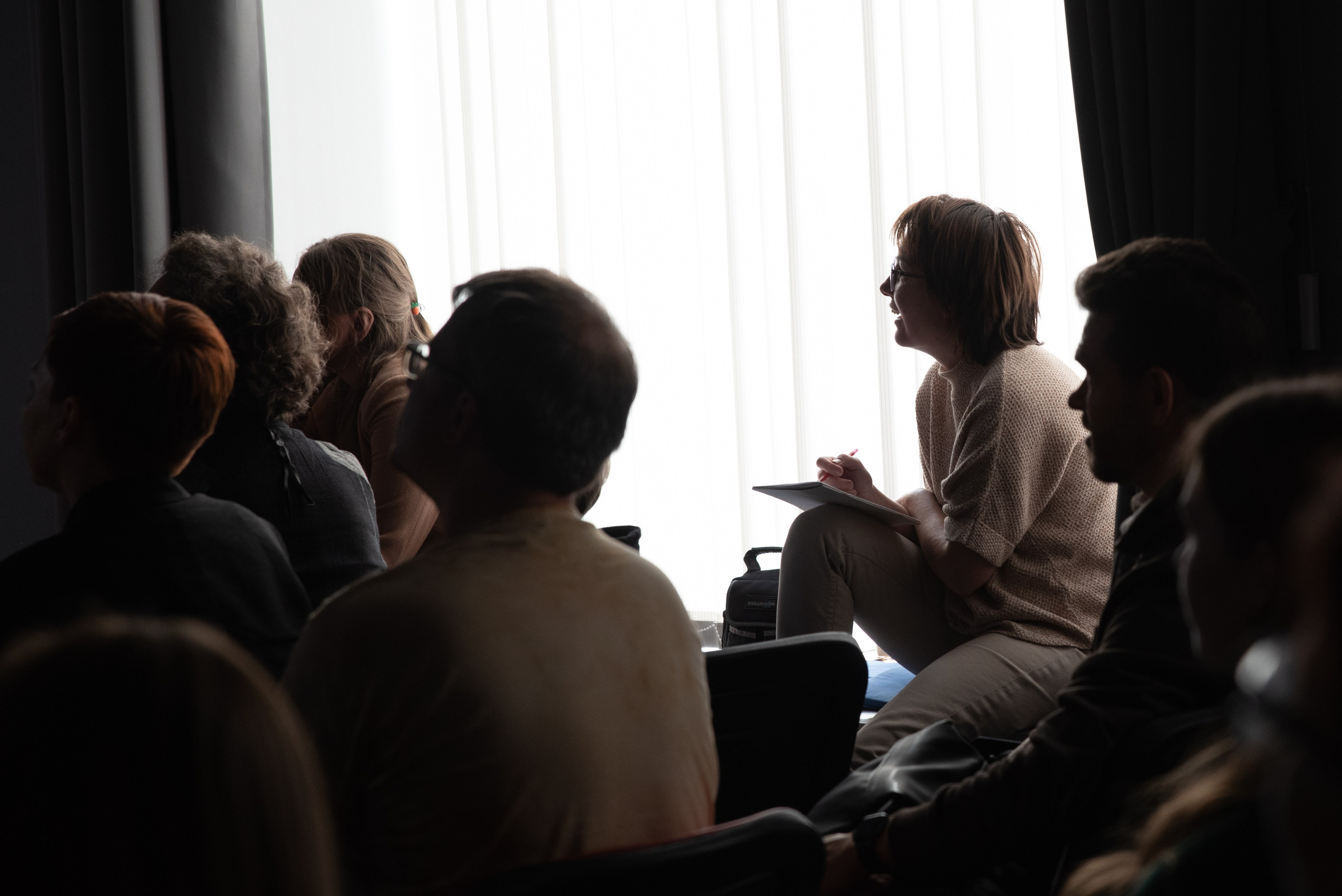 Женщина на лекции в фотошколе. Сидит у окна, держит блокнот и ручку, смеётся.