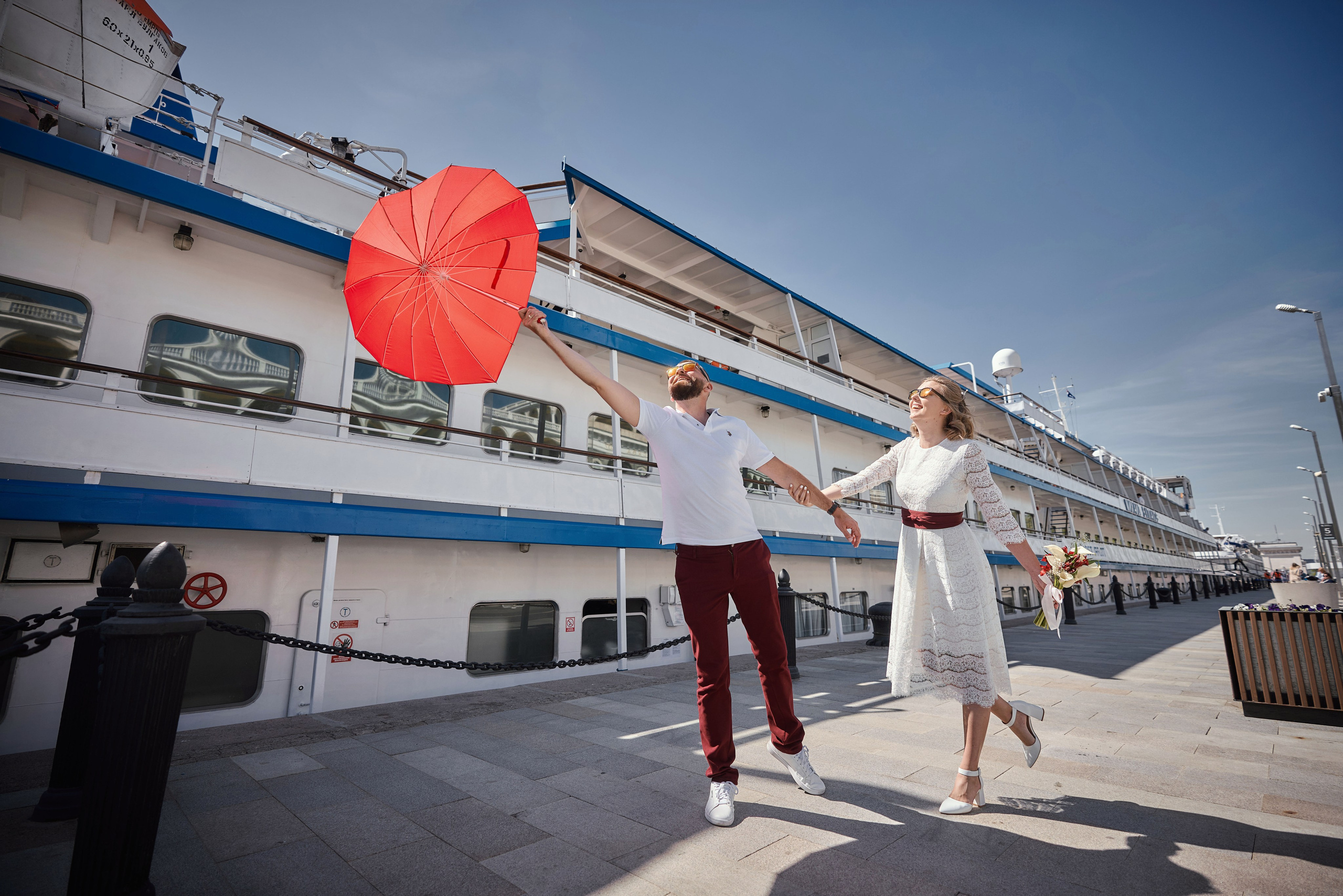 с девушкой идет мужчина который держится за улетающий красный зонт в форме сердца на причале речного вокзала на фоне речного корабля