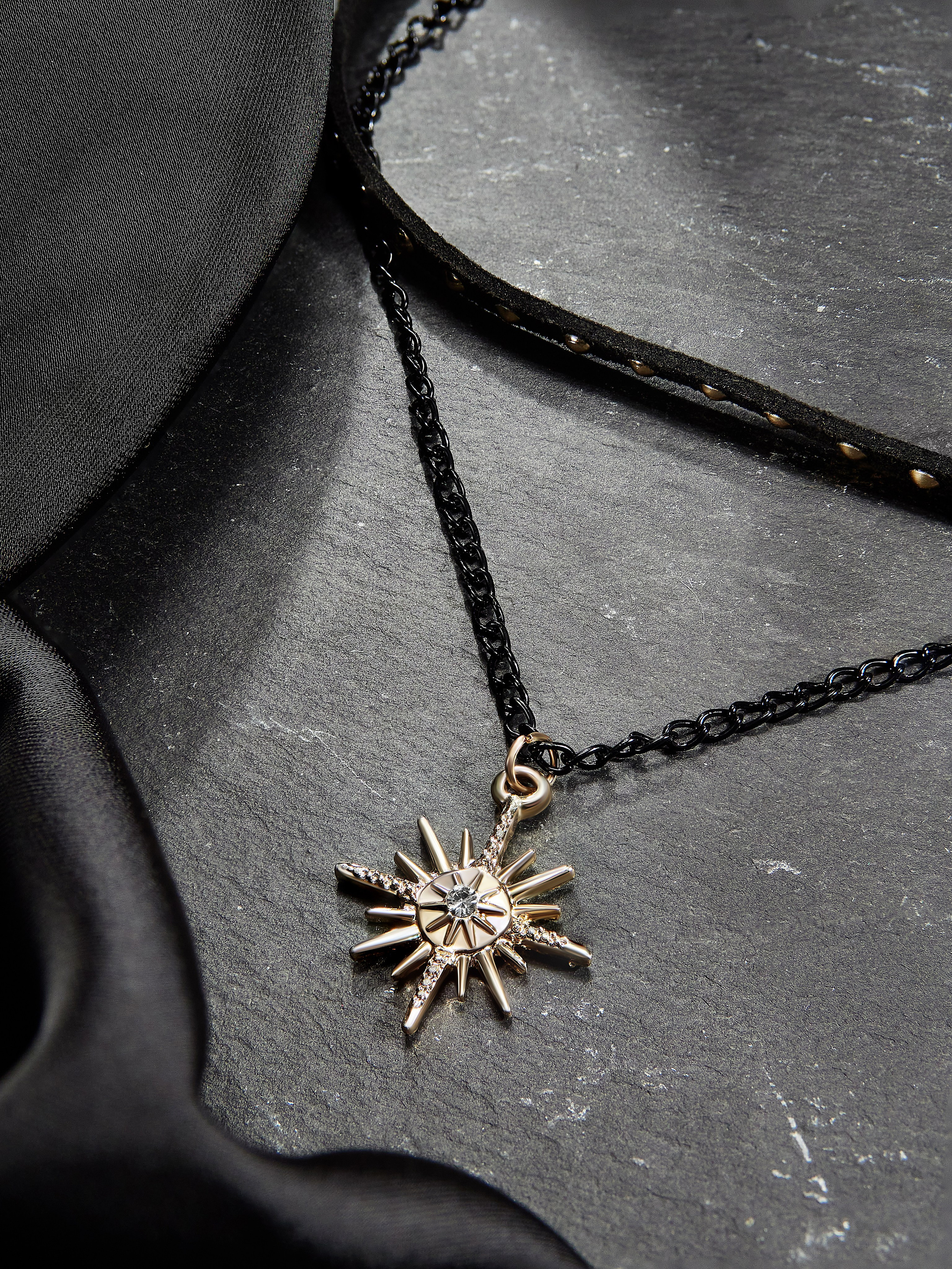 Стильное колье-цепочка черного цвета с подвеской в ​​форме звезды с замысловатыми деталями и небольшим драгоценным камнем в центре. Ожерелье расположено на темном сланце, рядом с которым драпирована мягкая ткань