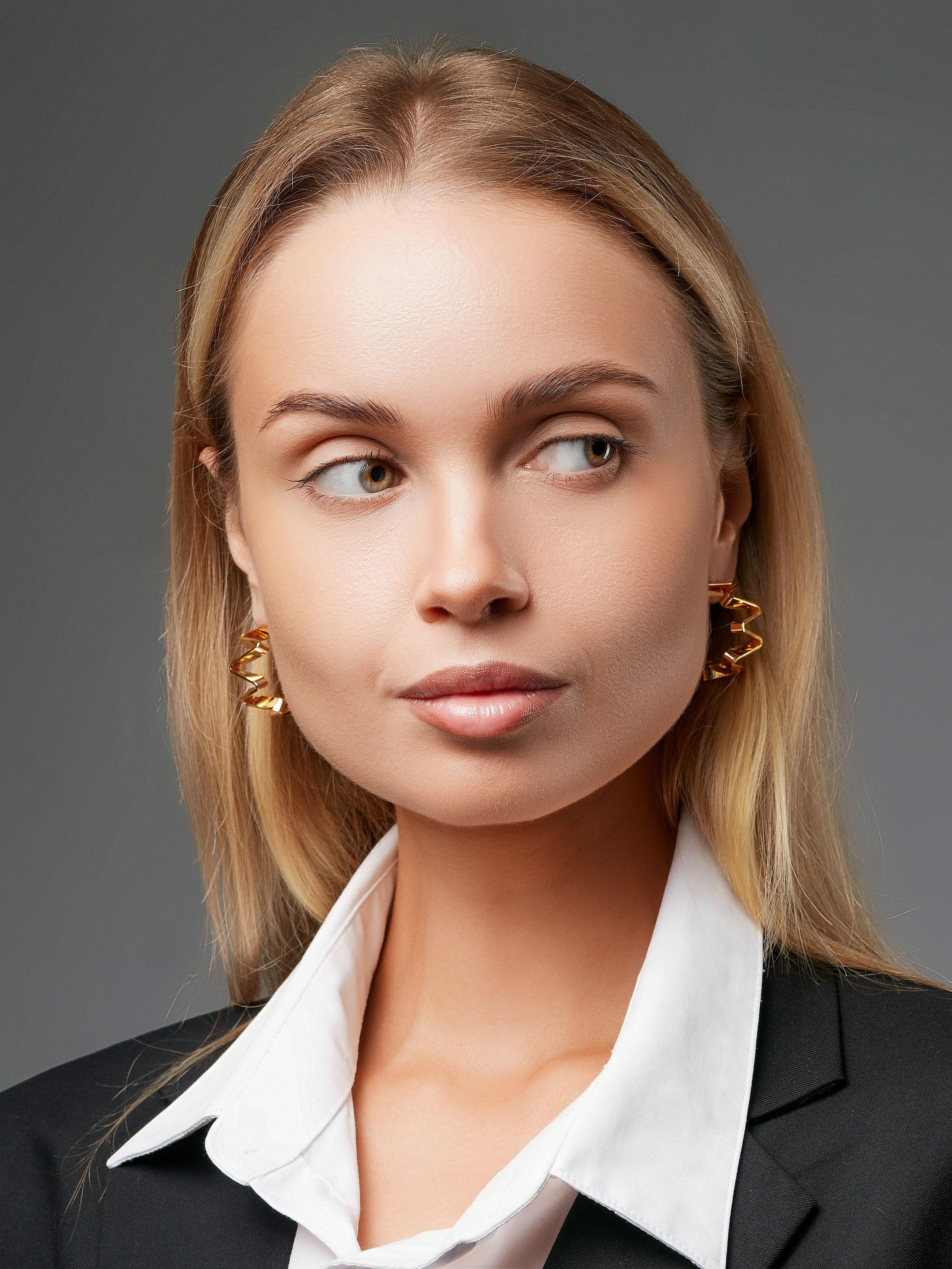 Женщина с длинными светлыми волосами, одетая в черный пиджак, белую рубашку и серьги-конго, смотрит направо. У нее золотые серьги и нейтральное выражение лица