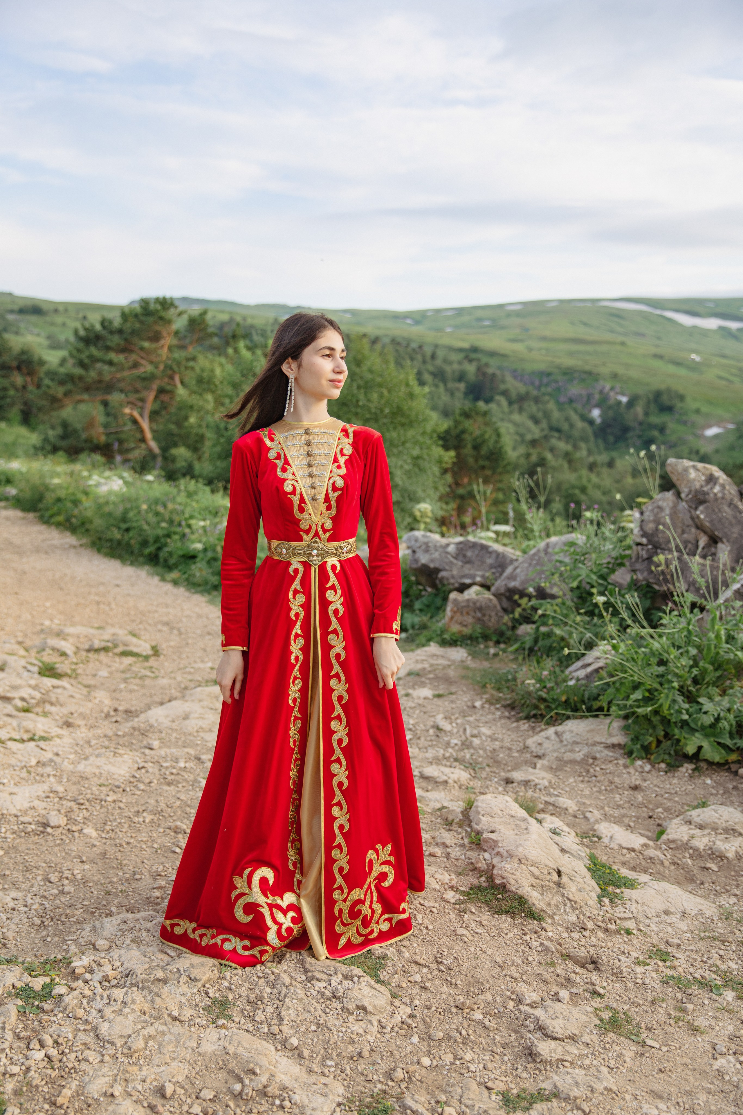 Красивая молодая девушка в традиционном адыгейском наряде, стоящая перед величественными горами.