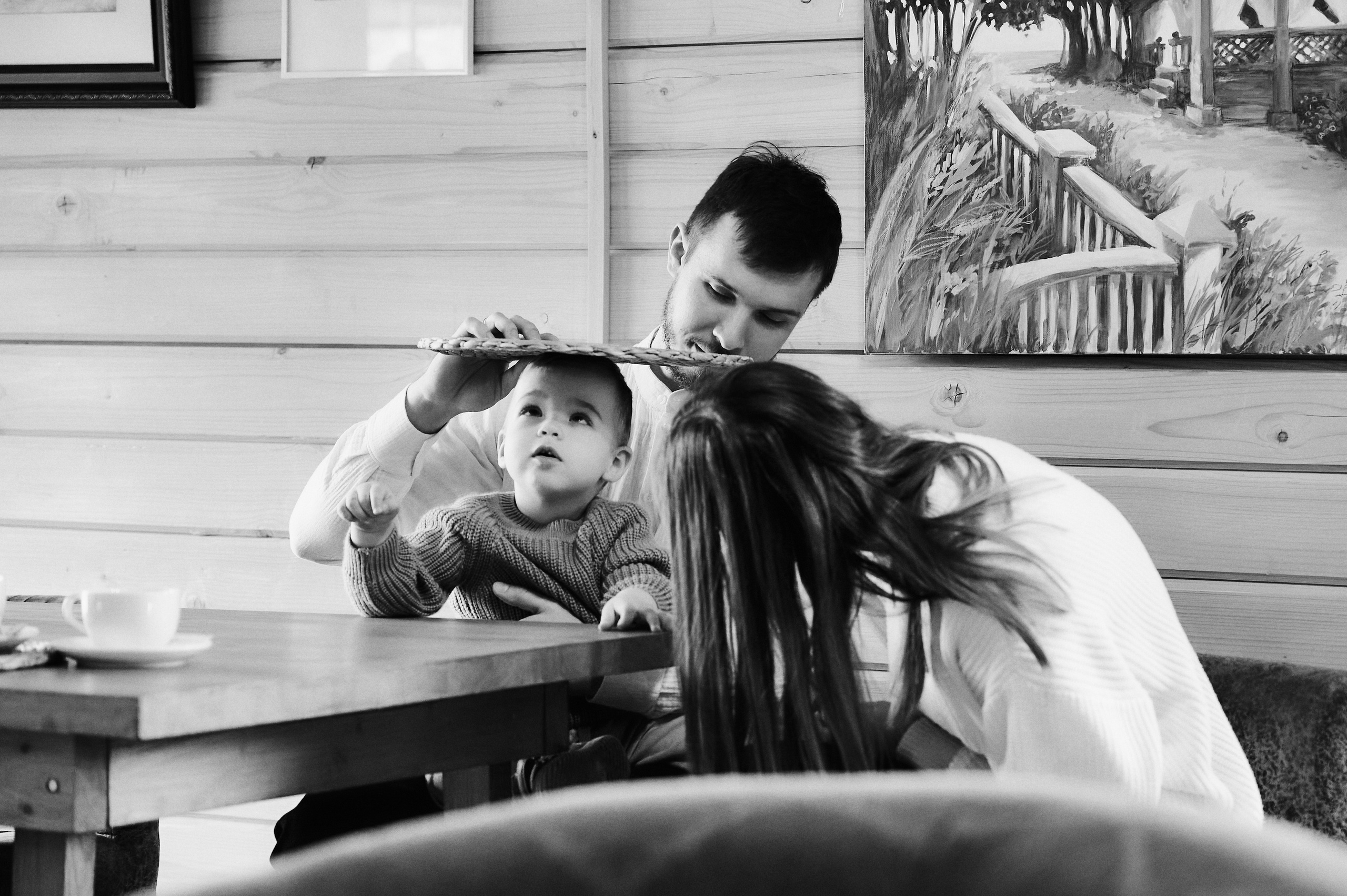 Отец играет с ребёнком, накрывая голову плетёной подставкой, мать смеётся рядом