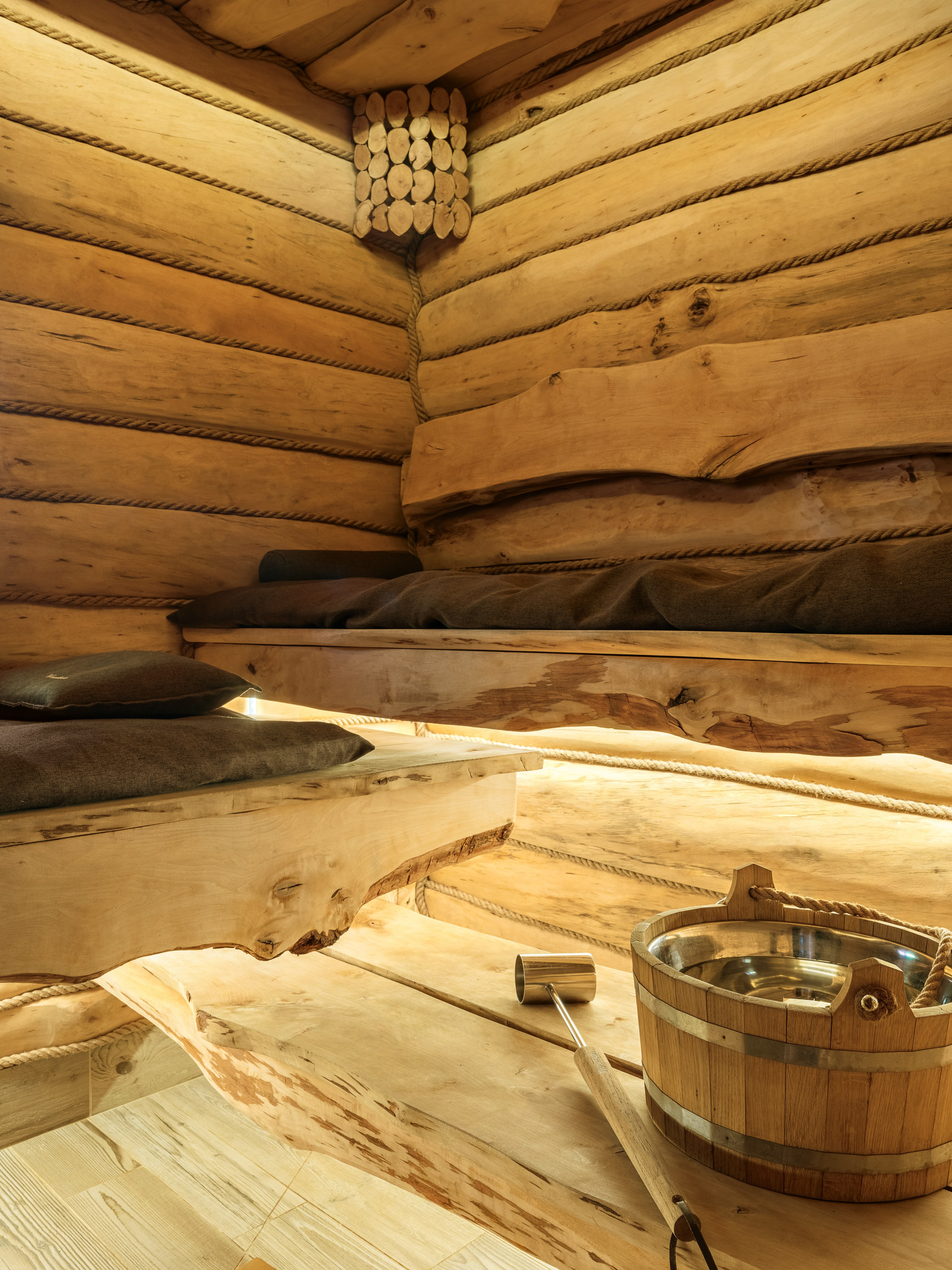 Интерьер традиционной деревянной сауны с полками из натурального бруса, ковшом и подушками, созданный дизайнером Наталией Шелепниковой, стилистом Софией Калбазовой и детально переданный фотографом Сергеем Денисенко в тёплом освещении.