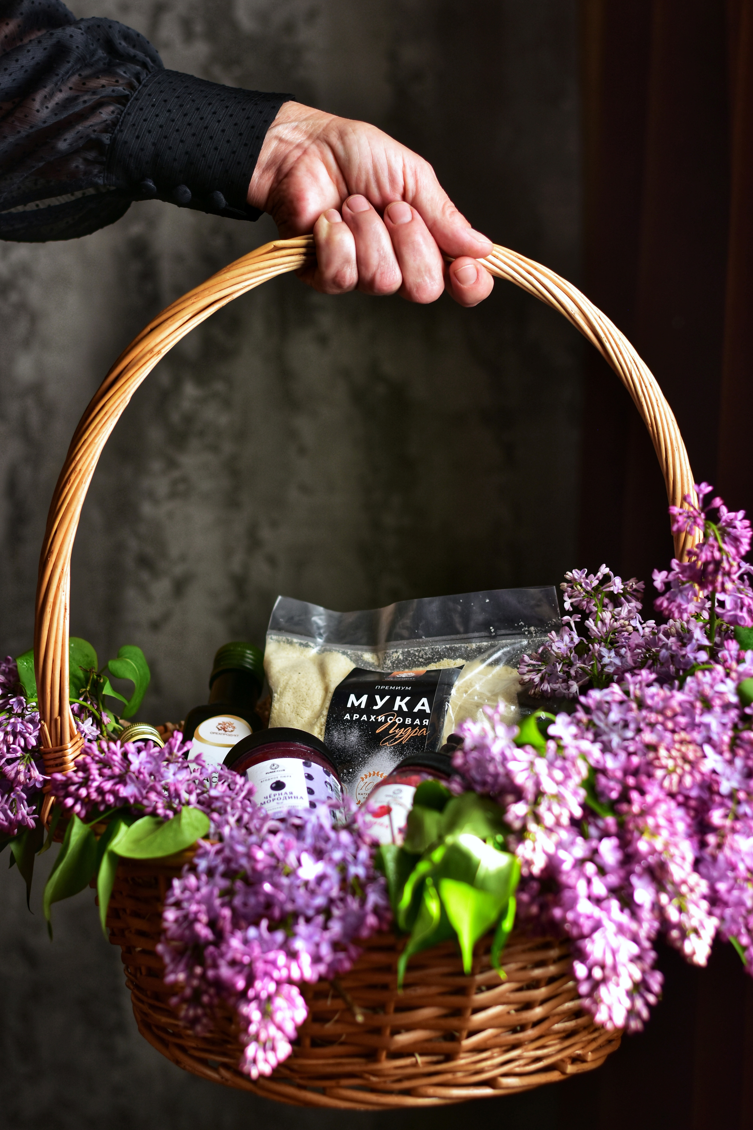 женская рука держит плетеную корзину, наполненную фиолетовыми цветами.