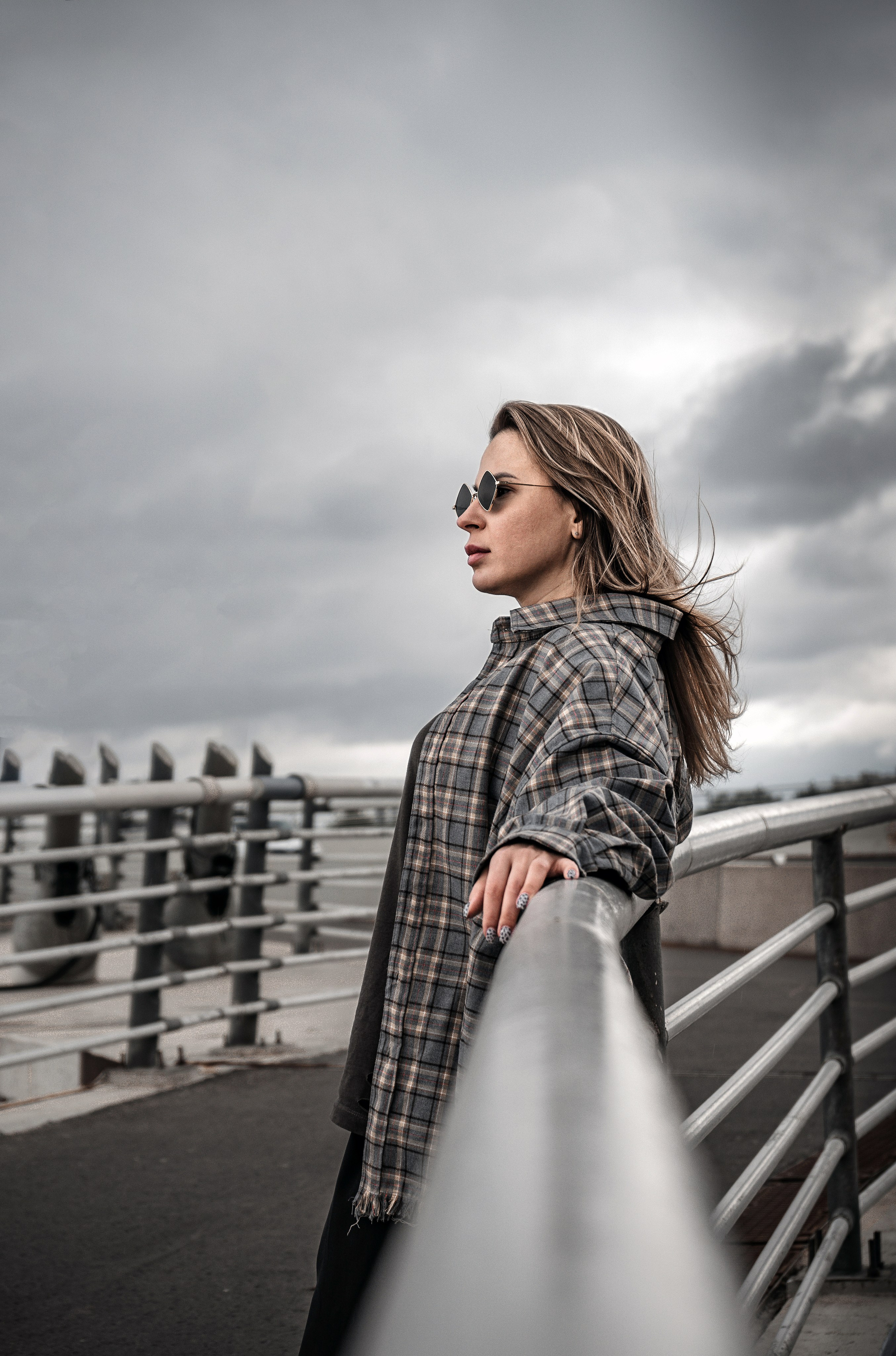 Индустриальный street style: девушка в клетчатой oversize-рубашке и широких чёрных брюках стоит боком на мосту, смотрит в даль, cat-eye солнцезащитные очки, развевающиеся волосы, тёмный урбан-бэкграунд.