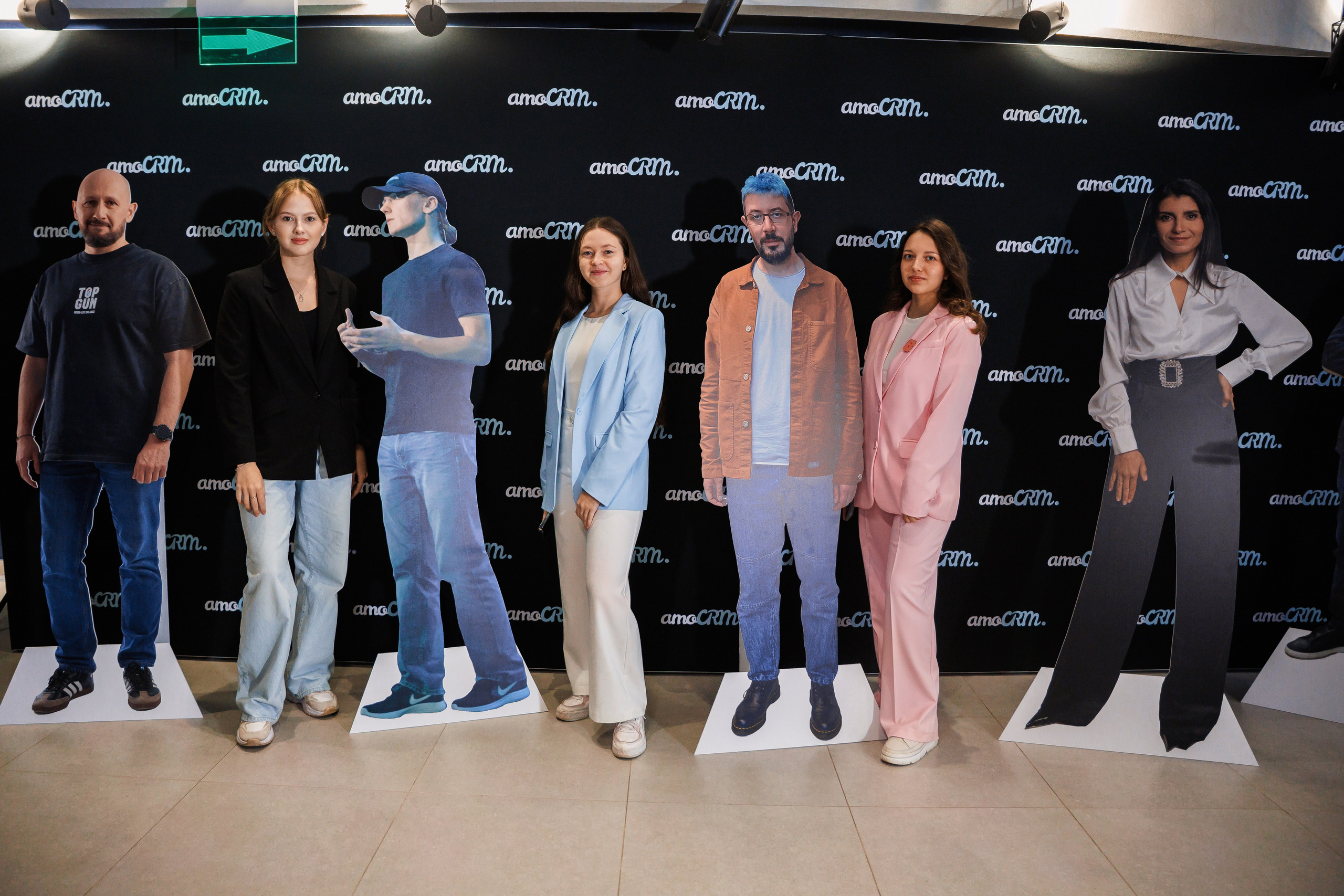 Шесть человек стоят в помещении рядом с картонными фигурами в натуральную величину, изображающими их самих, все улыбаются, на фоне черной стены с повторяющимися логотипами amaCRM. Группа одета в повседневную и деловую одежду.