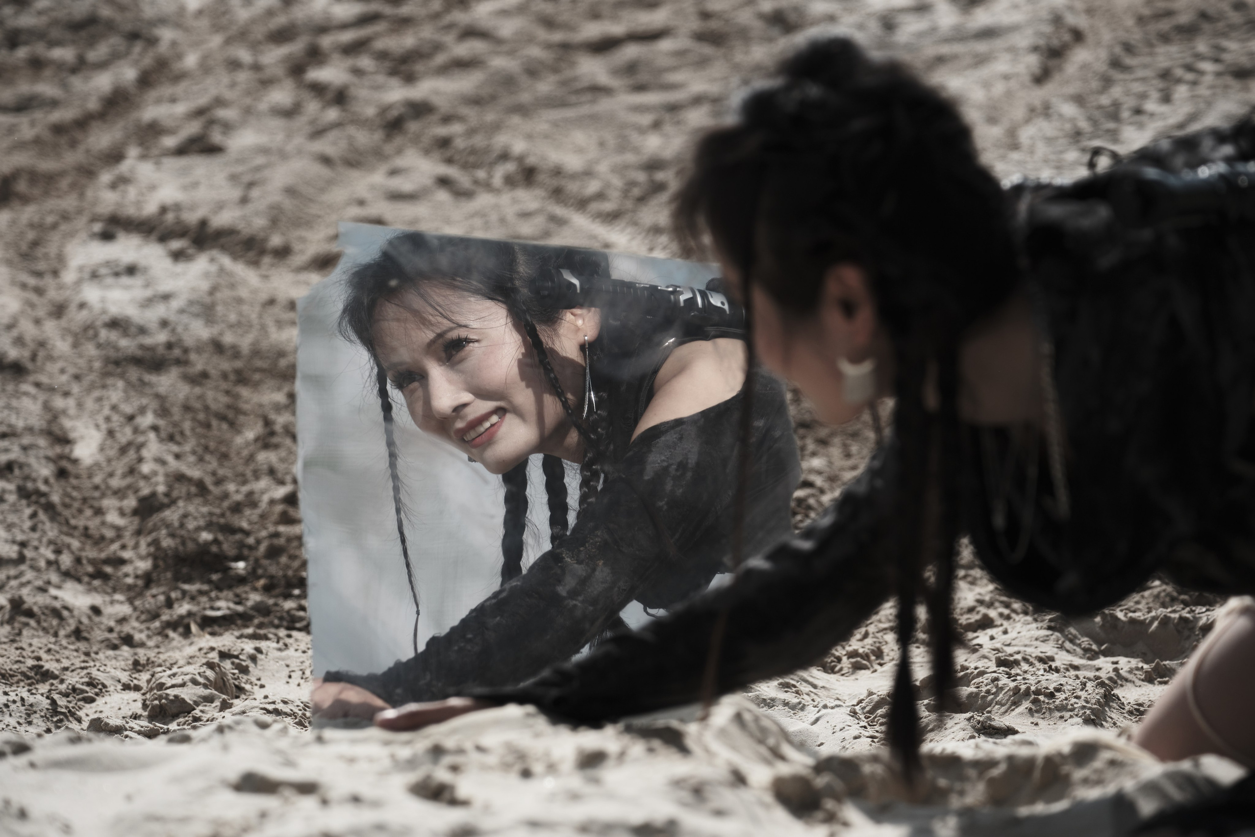 Девушка азиатской внешности ползет по песку и смотрит в зеркало