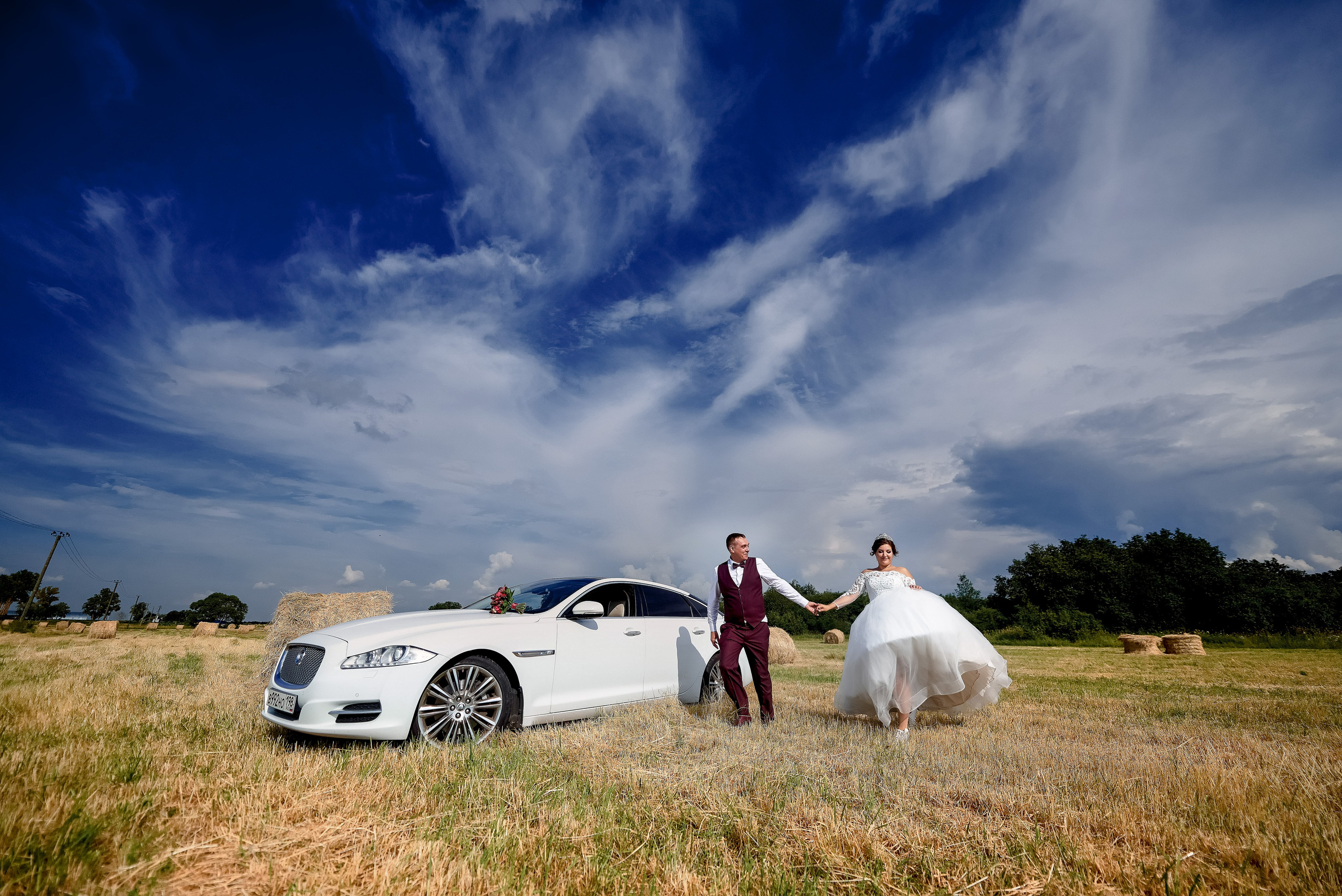 Свадебная пара бежит по бескрайнему полю рядом со свадебным автомобилем на фоне прекрасного голубого неба