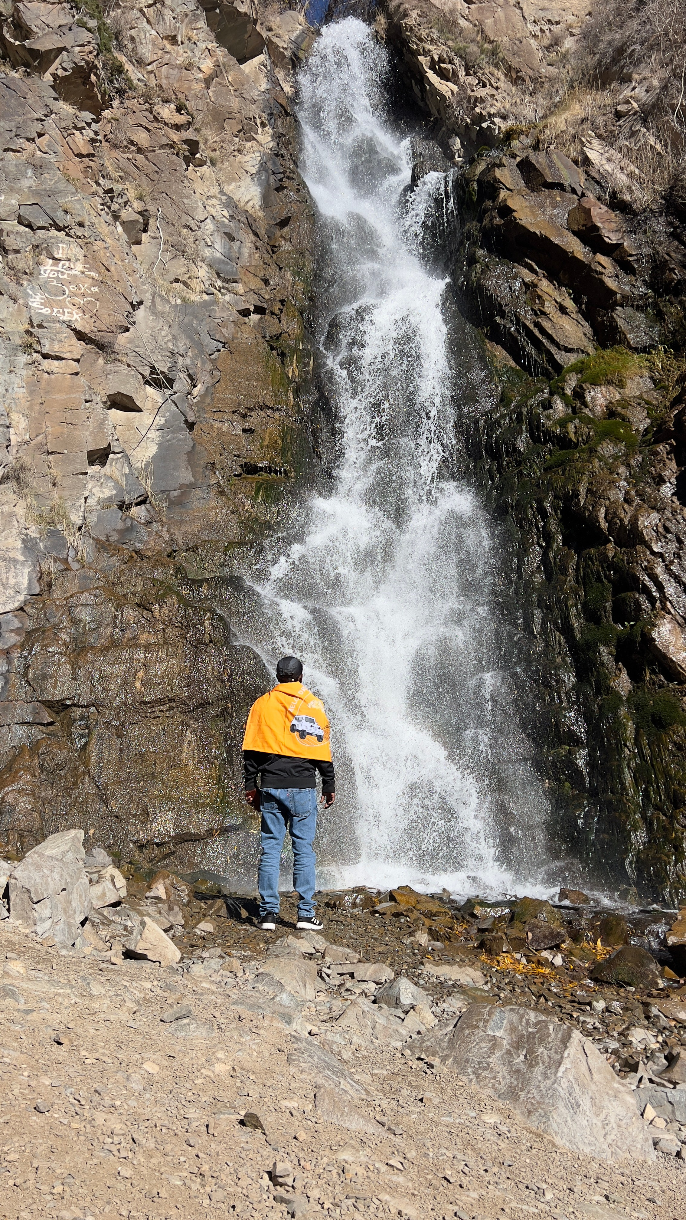 Человек стоит у подножия водопада, одетый в яркую оранжевую накидку или флаг с изображением автомобиля и логотипа агентства путешествий Melon Drive. Водопад Медвежий падает с высоты по скалистой поверхности, окружённой камнями и мхом. Чистая вода струится вниз, создавая естественный фон. Местность вокруг сухая и каменистая, с ясным солнечным освещением, подчеркивающим детали природы.