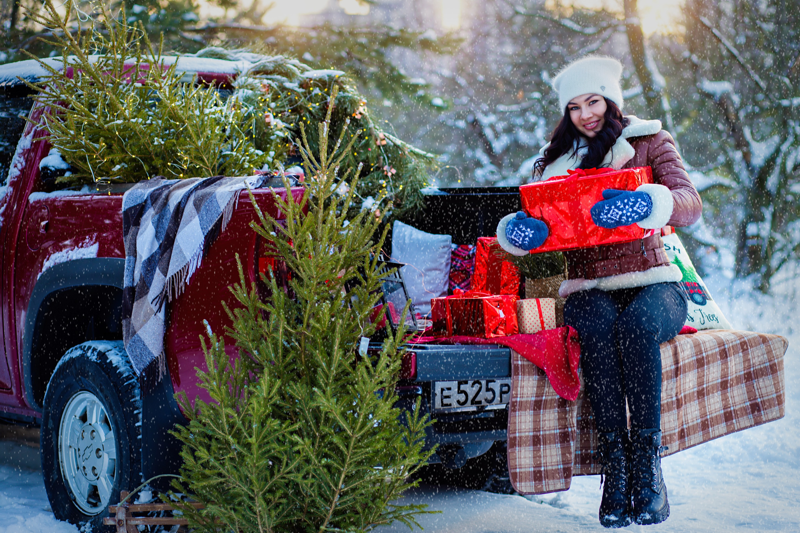 девушка в кожаной  куртке коричневого  цвета    сидит  на   пледе  в  клетку   в машине  пикап красного цвета. у нее в  руках красный подарок. вокруг елки. снег. зима.