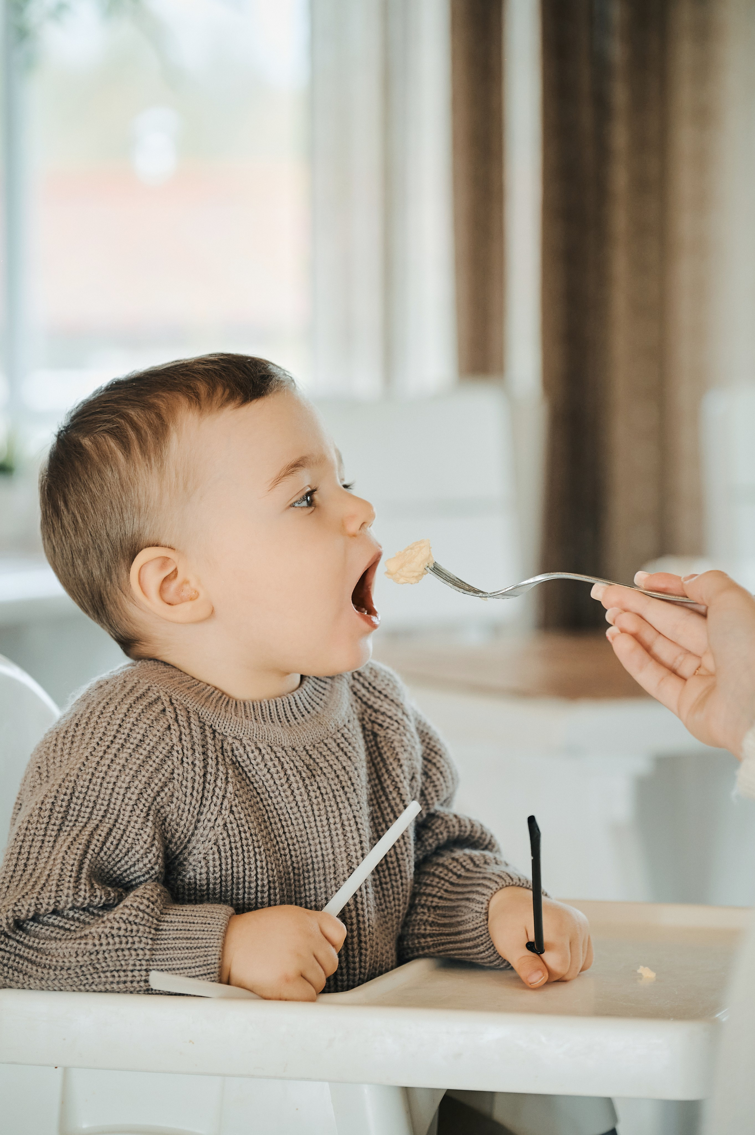 Мальчик широко открывает рот, готовясь съесть ложку еды, поднесённую взрослым.