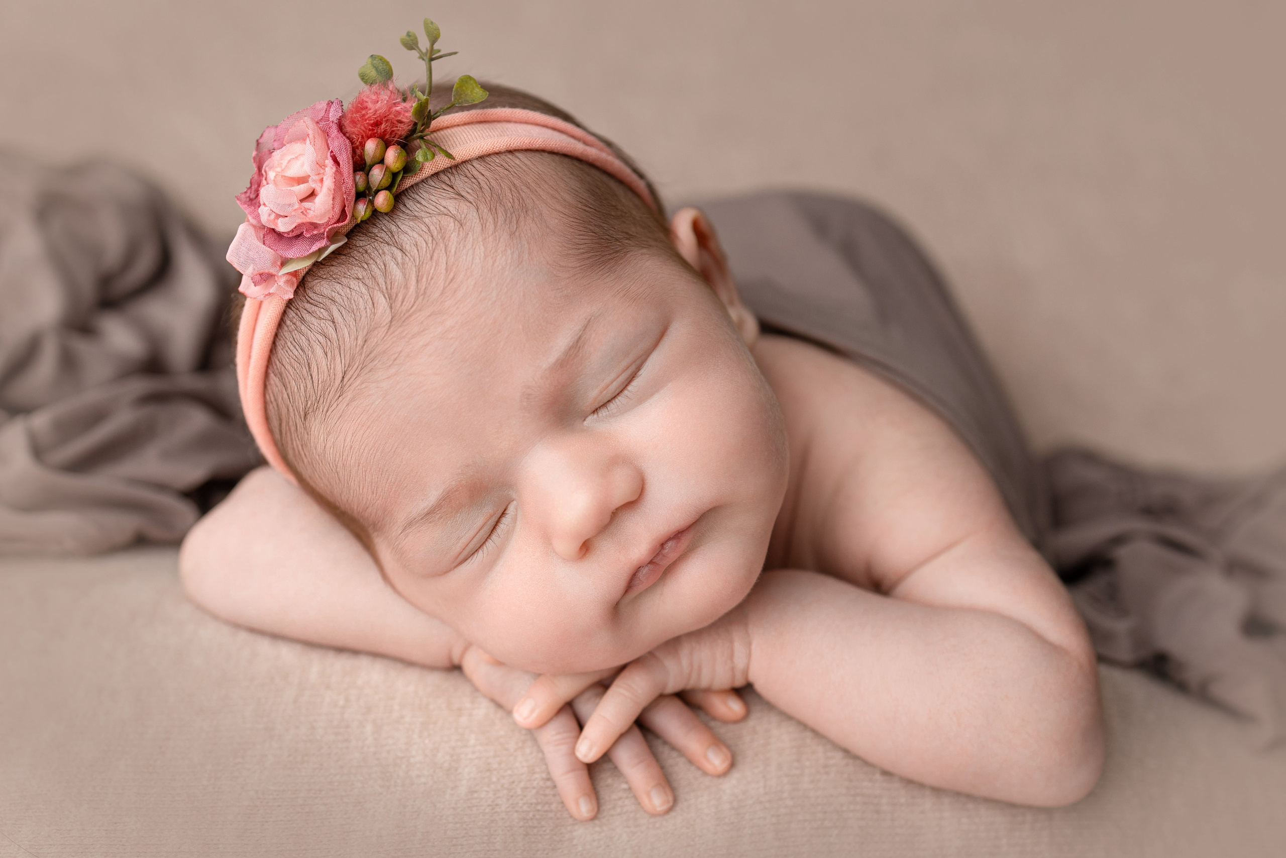 Спящий новорожденный с розовым цветочным ободком, уложенный на сером фоне.