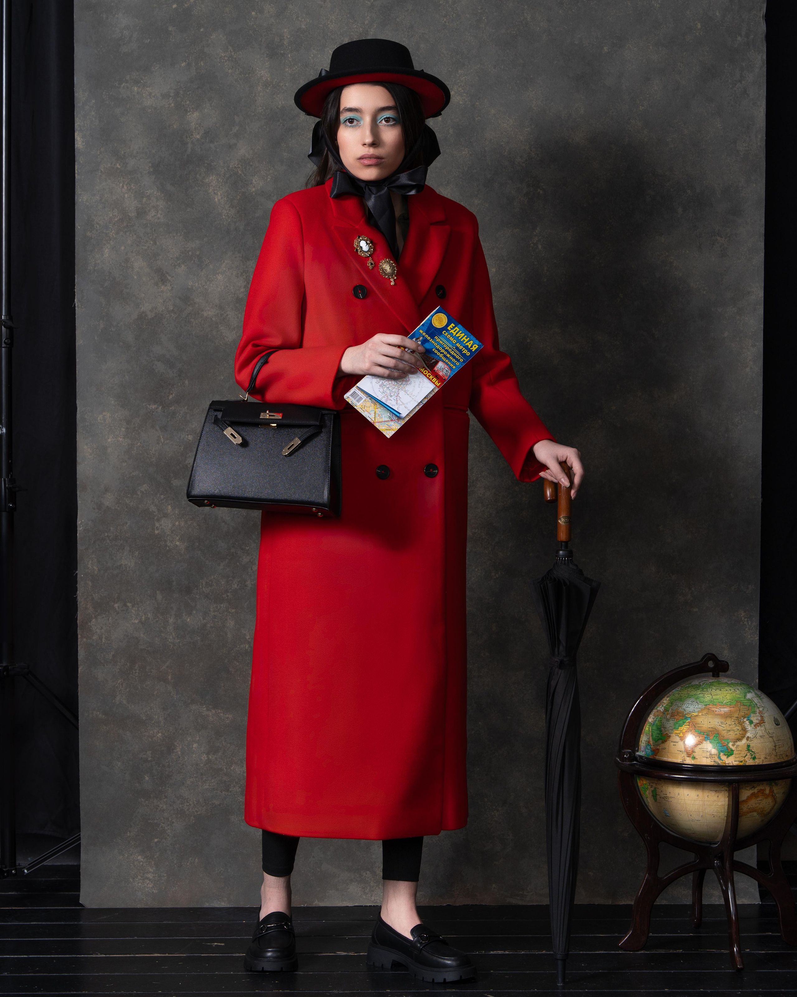 На фотографии изображена женщина, одетая в красное пальто и черную шляпу с бантом. Она стоит на фоне серой стены, которая создает контраст с ее ярким нарядом. В одной руке она держит черный зонт, а в другой — журнал "Европа Плюс". На поясе пальто виден декоративный элемент в виде цветка. Женщина также носит черную сумку через плечо и черные туфли. Ее взгляд направлен прямо на камеру, а выражение лица серьезное и сосредоточенное.