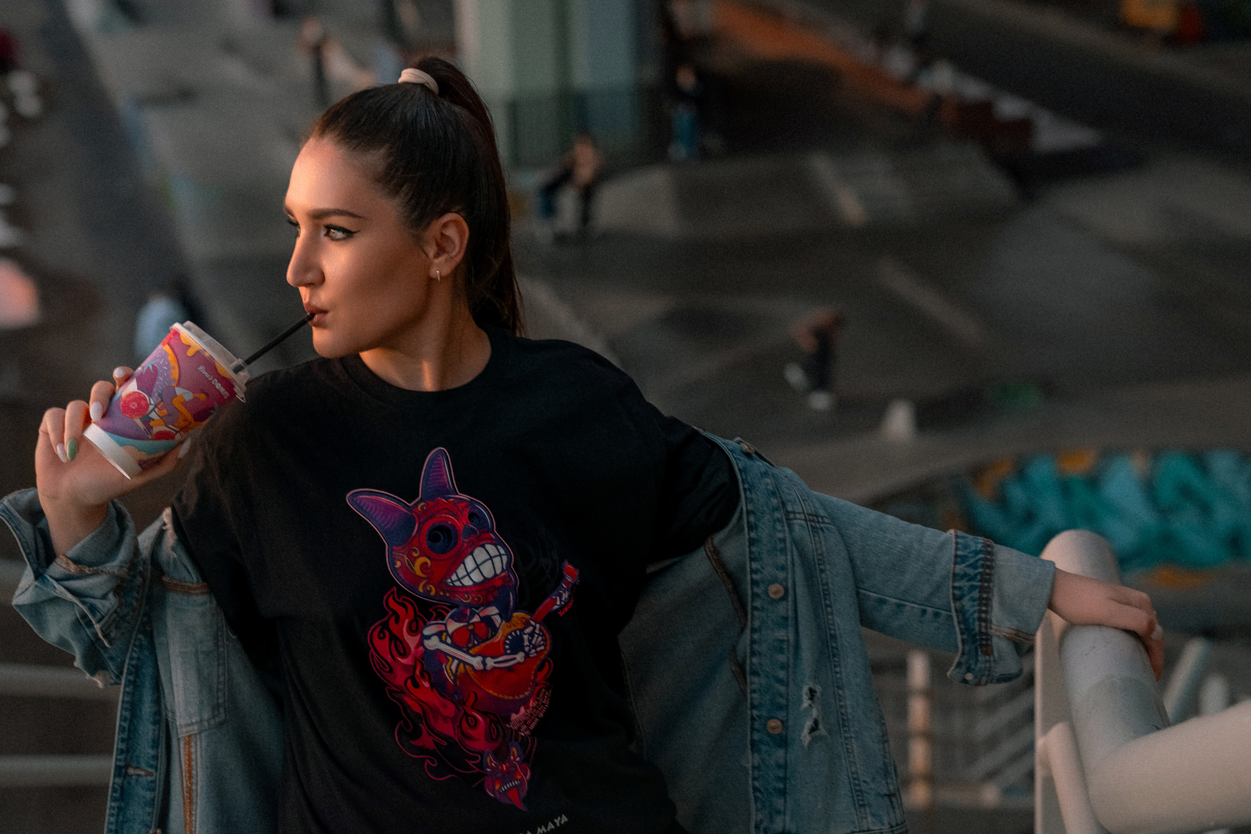 Девушка в джинсовой куртке пьет кофе на фоне скейт-площадки и рамп