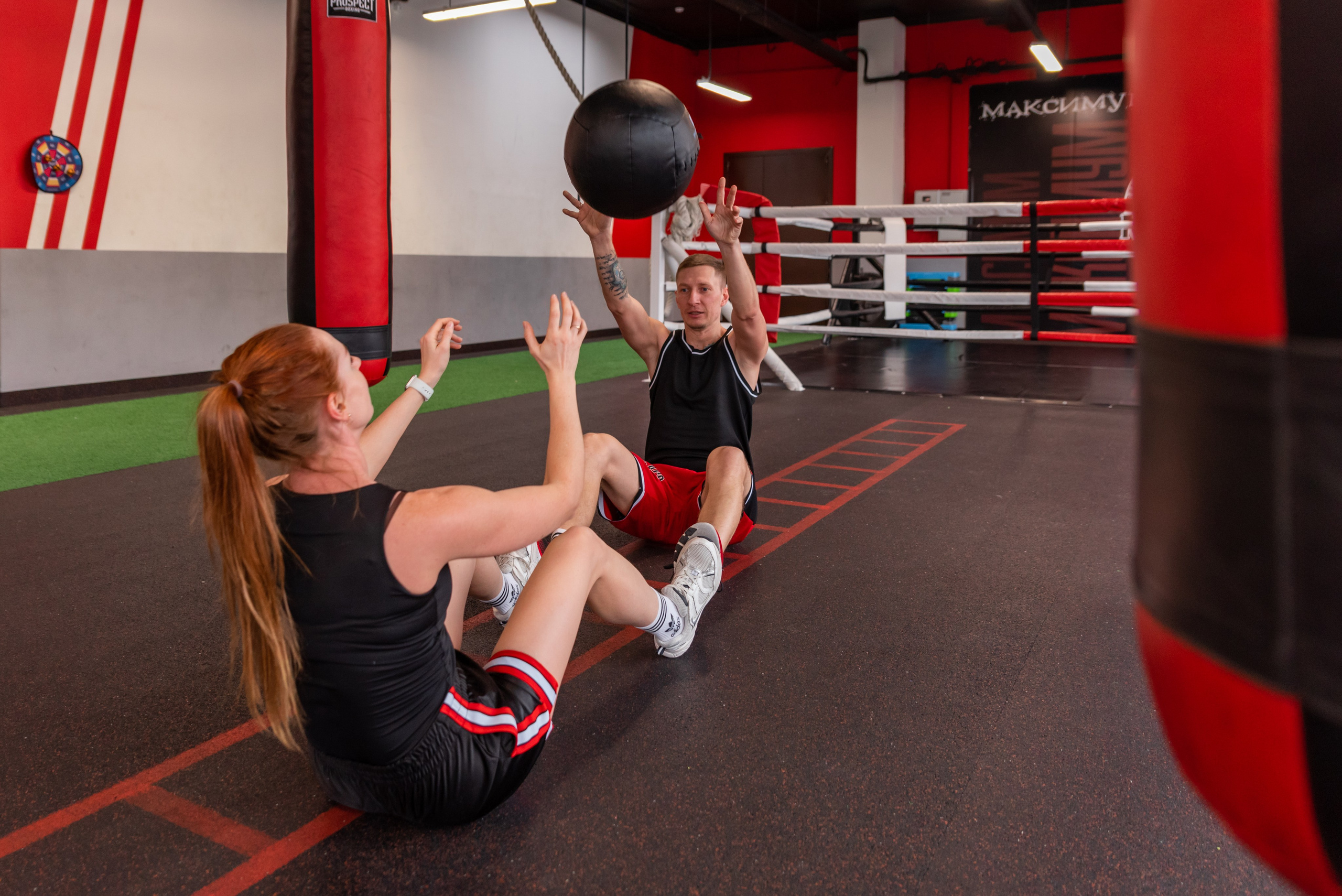 Молодая женщина и мужчина сидят на полу в спортзале и перекидывают друг другу тяжелый мяч