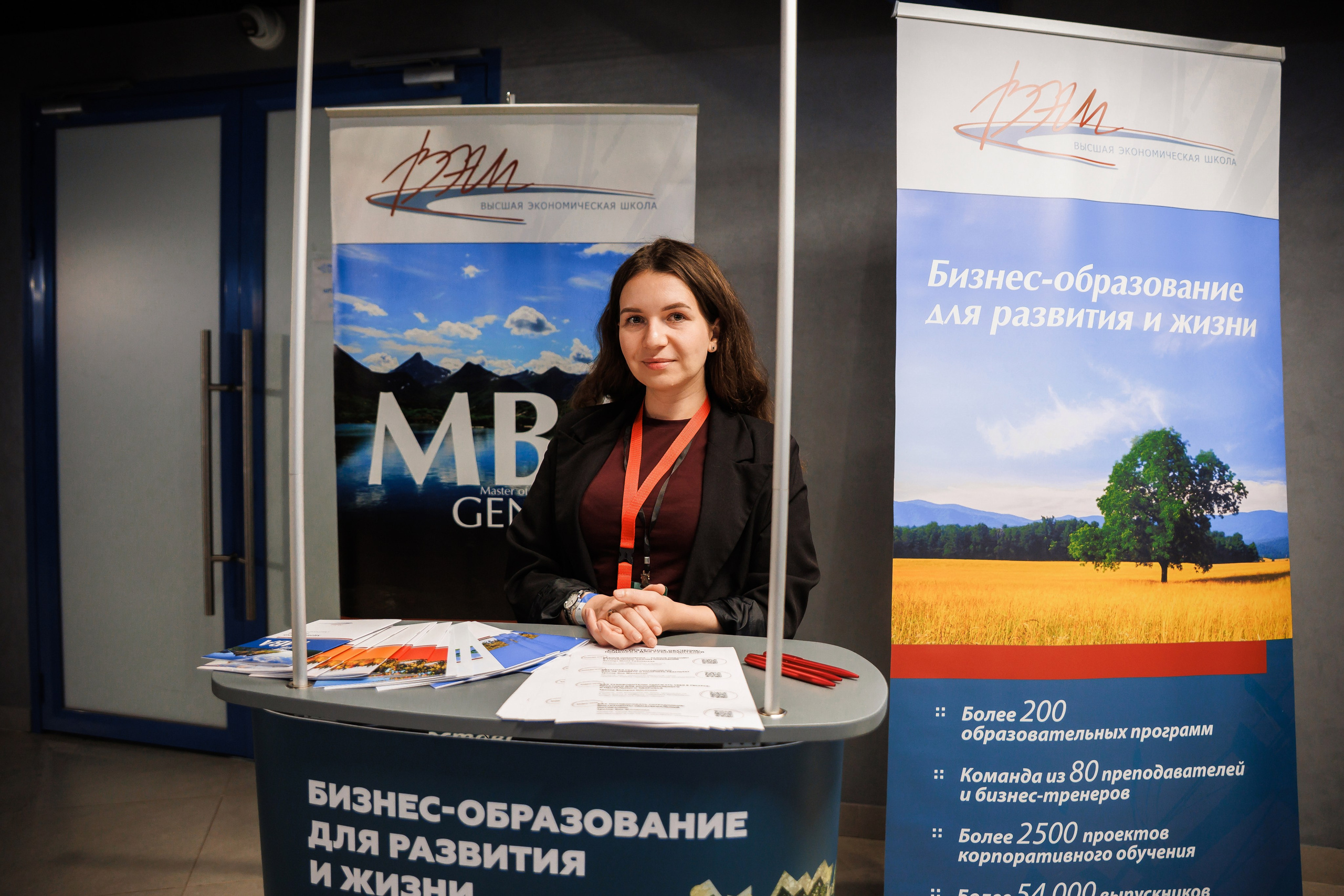 Женщина стоит за рекламным стендом с брошюрами и двумя баннерами о бизнес-образовании на русском языке. На баннерах изображены живописные пейзажи и информация о программах MBA.