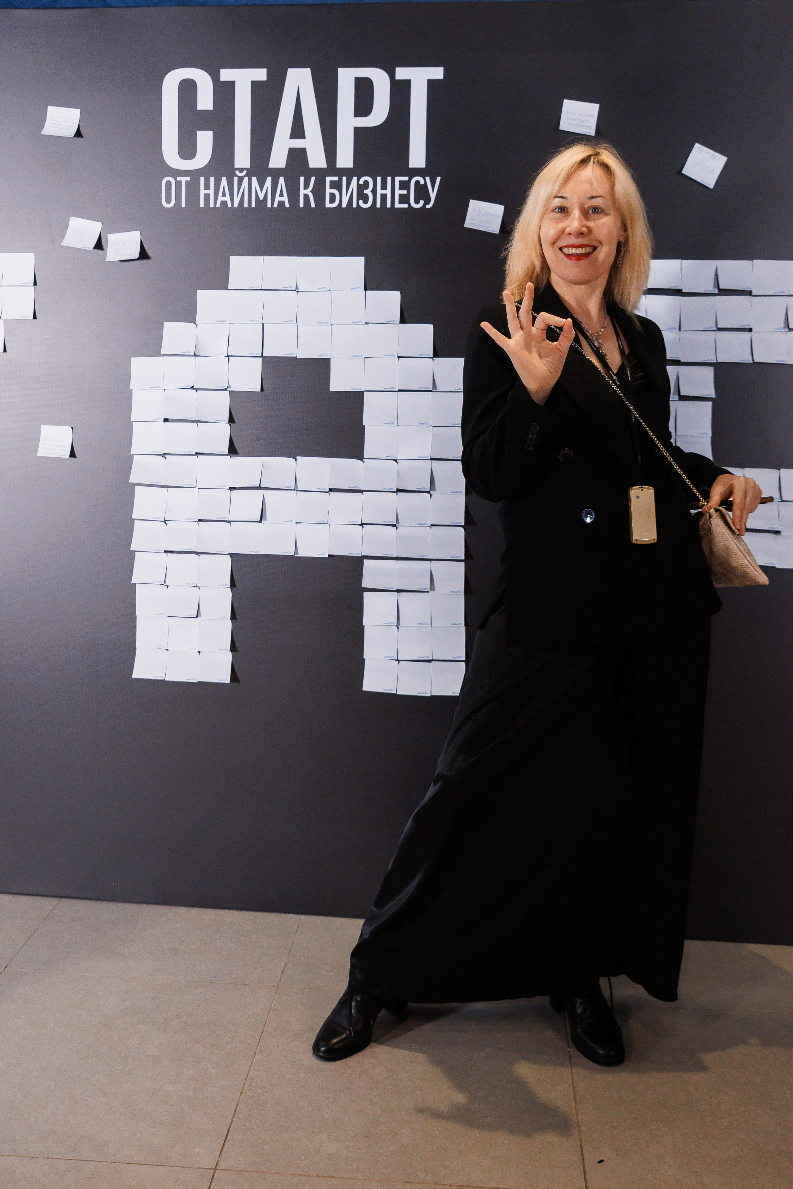 Улыбающаяся женщина в черной одежде позирует на фоне стены с надписями на русском языке и рисунком из стикеров, показывая жестом «ОК».