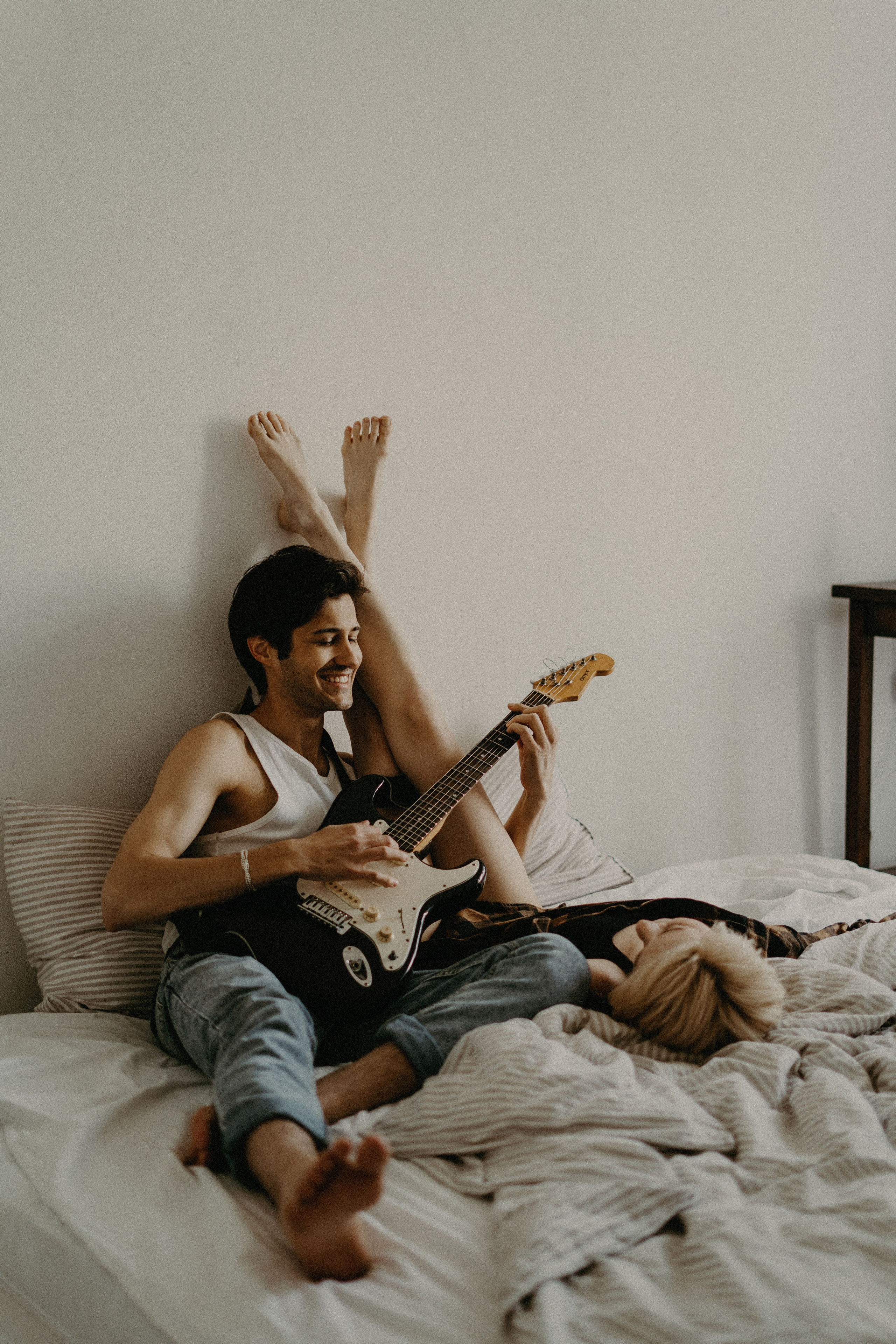 Молодой мужчина в джинсах лежит на кровати с гитарой и поет для девушки, которая лежит рядом