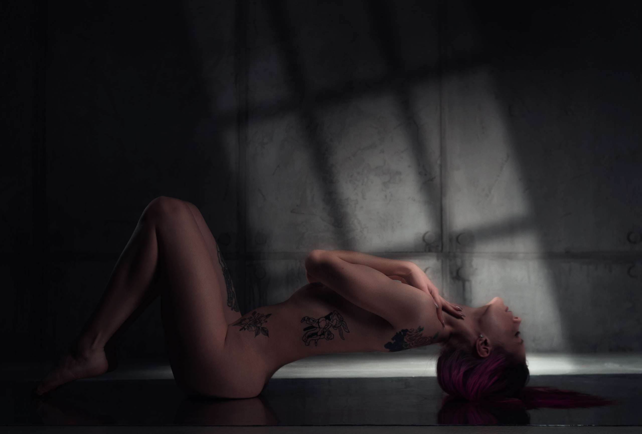 Фотосессия ню в низком ключе. Девушка лежит на спине, на сером фоне тени от окна.