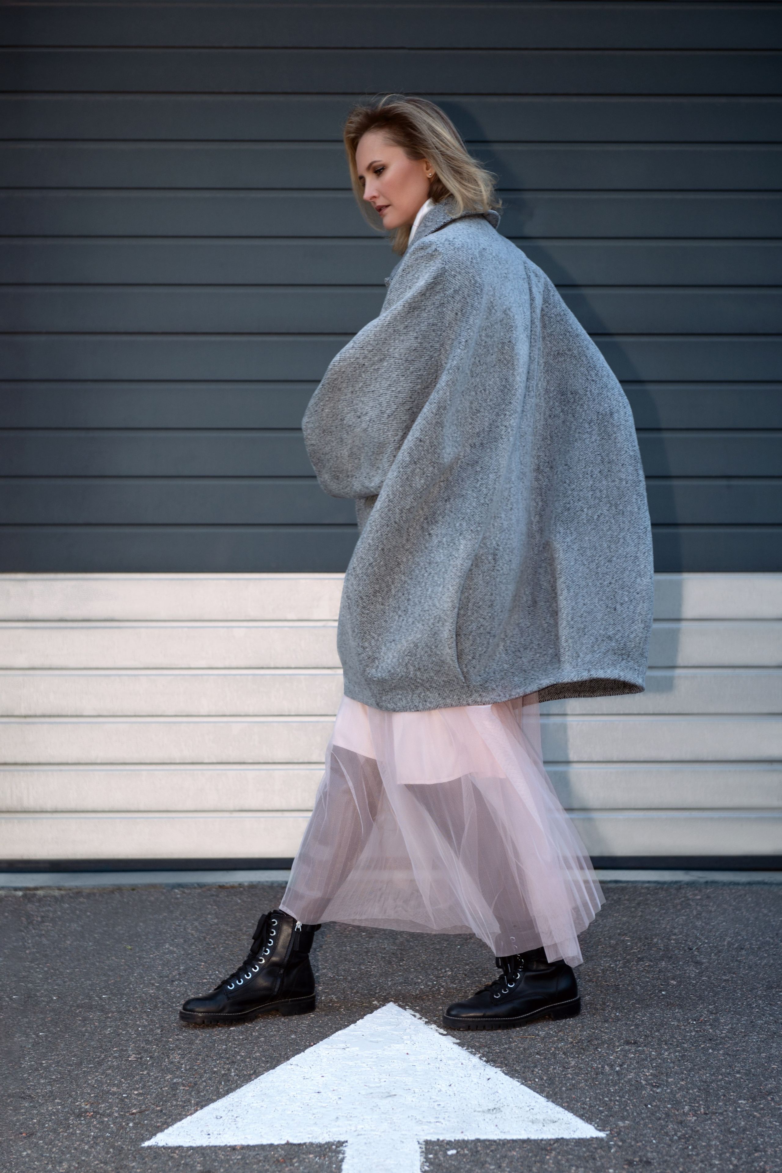 Женщина идет вдоль серой стены. На ней серое пальто-кокон, розовая юбка из фатина и черные грубые ботинки. Уличная контент-съемка для бутика. Съемка со смешанным светом. Вспышка + естественный свет