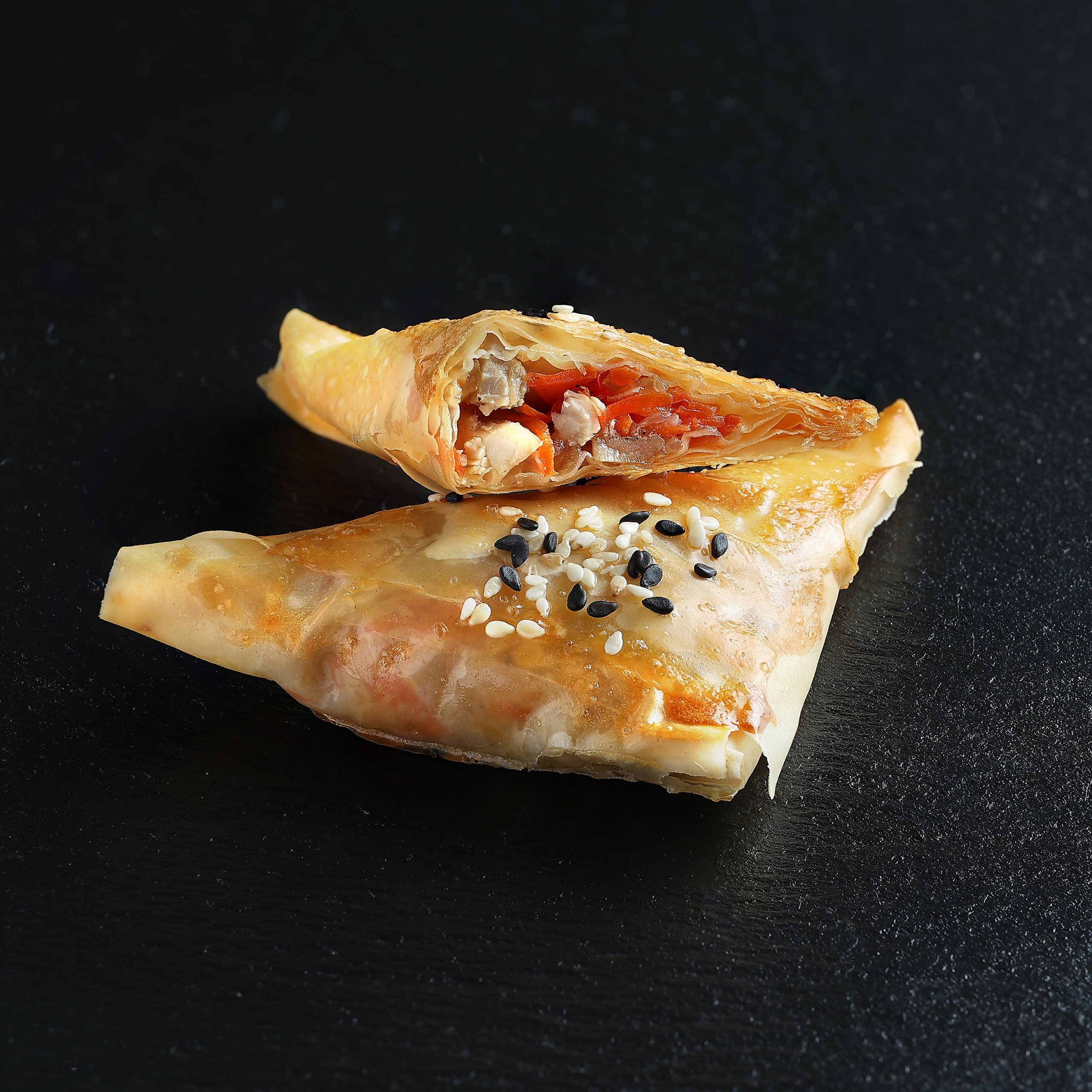 Слоёный растягай с рыбой, луком и болгарским перцем, украшенный белым и чёрным кунжутом, на тёмном фоне.