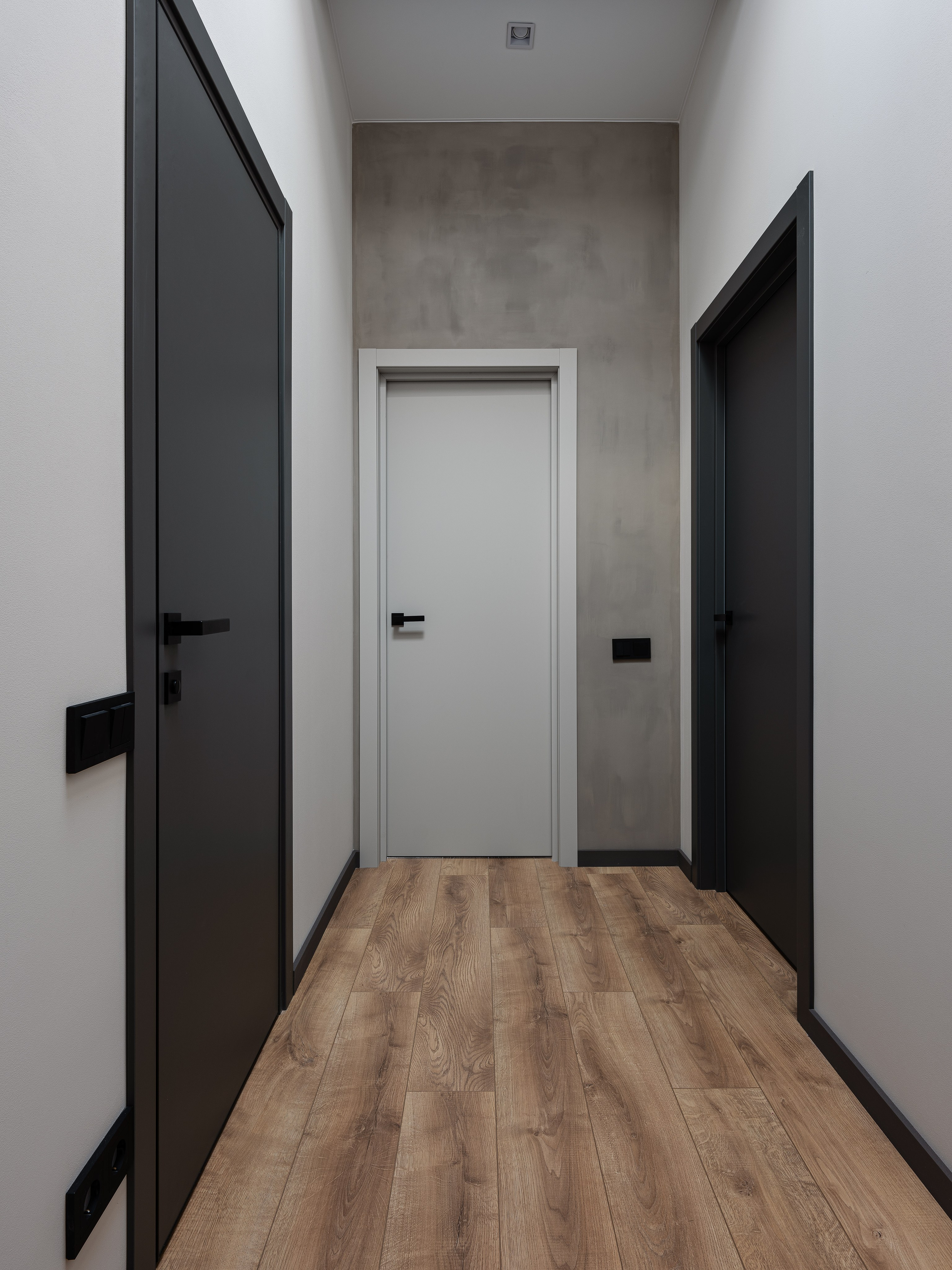 Интерьер коридора с серыми стенами и деревянным полом, чёрные двери создают выразительный контраст. Дизайн в минимализме от Екатерины Мищенко, фотография Сергея Денисенко, стильный современный жилой проект.