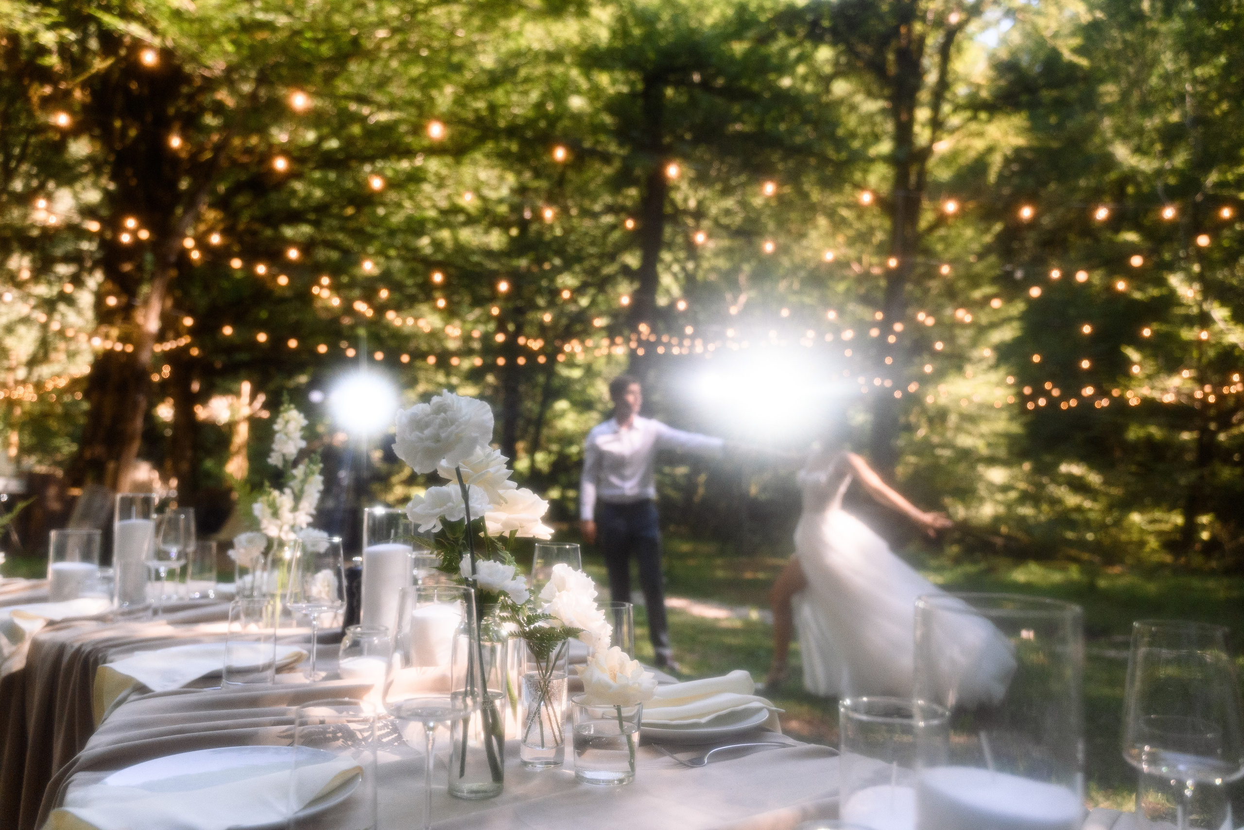 Красивый кадр в лесу на котором на переднем плане изображен стол накрытый на нём стоит посуда лежат салфетки сверху много-много гирлянд и где-то посередине кадра жених с невестой репетируют свой танец