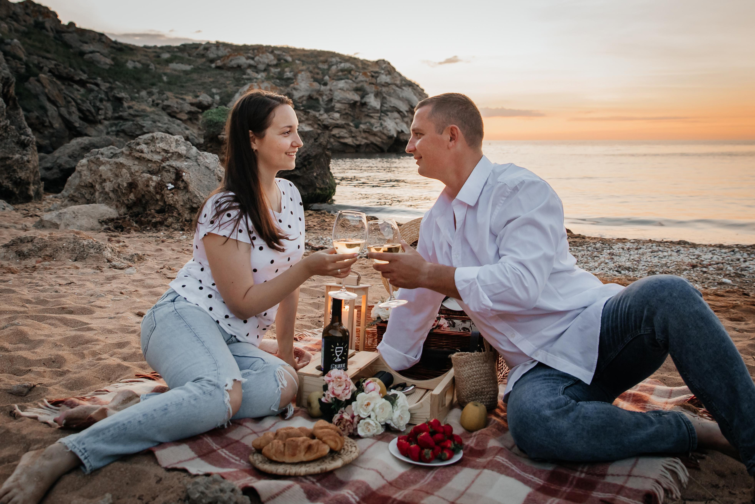 Влюбленная пара на берегу моря на закате сидят на пледе , у них пикник, пьют вино и смотрят друг на друга. 