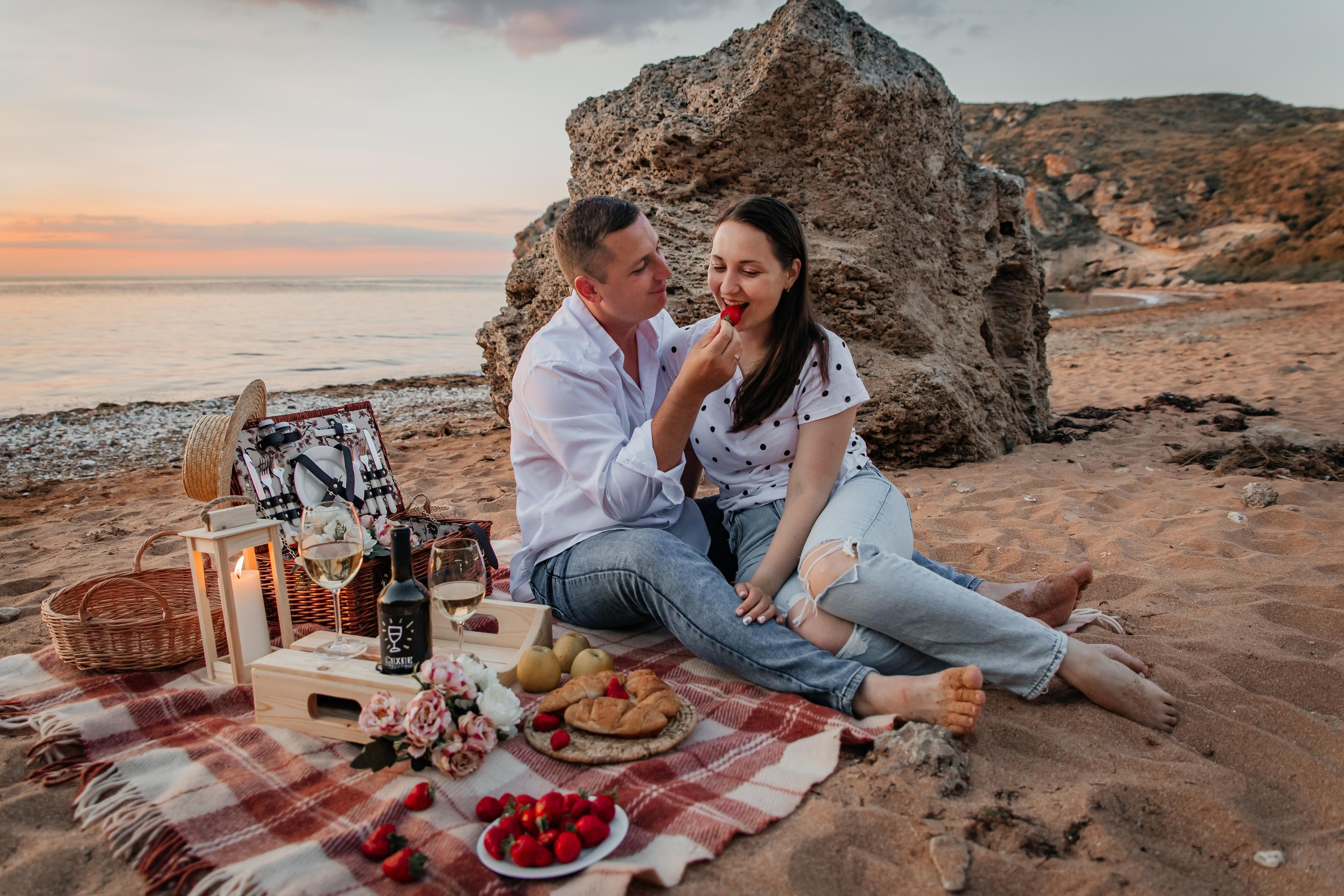 Пикник на морском побережье на закате влюбленная пара сидит на пледе , парень кормит девушку клубникой. 