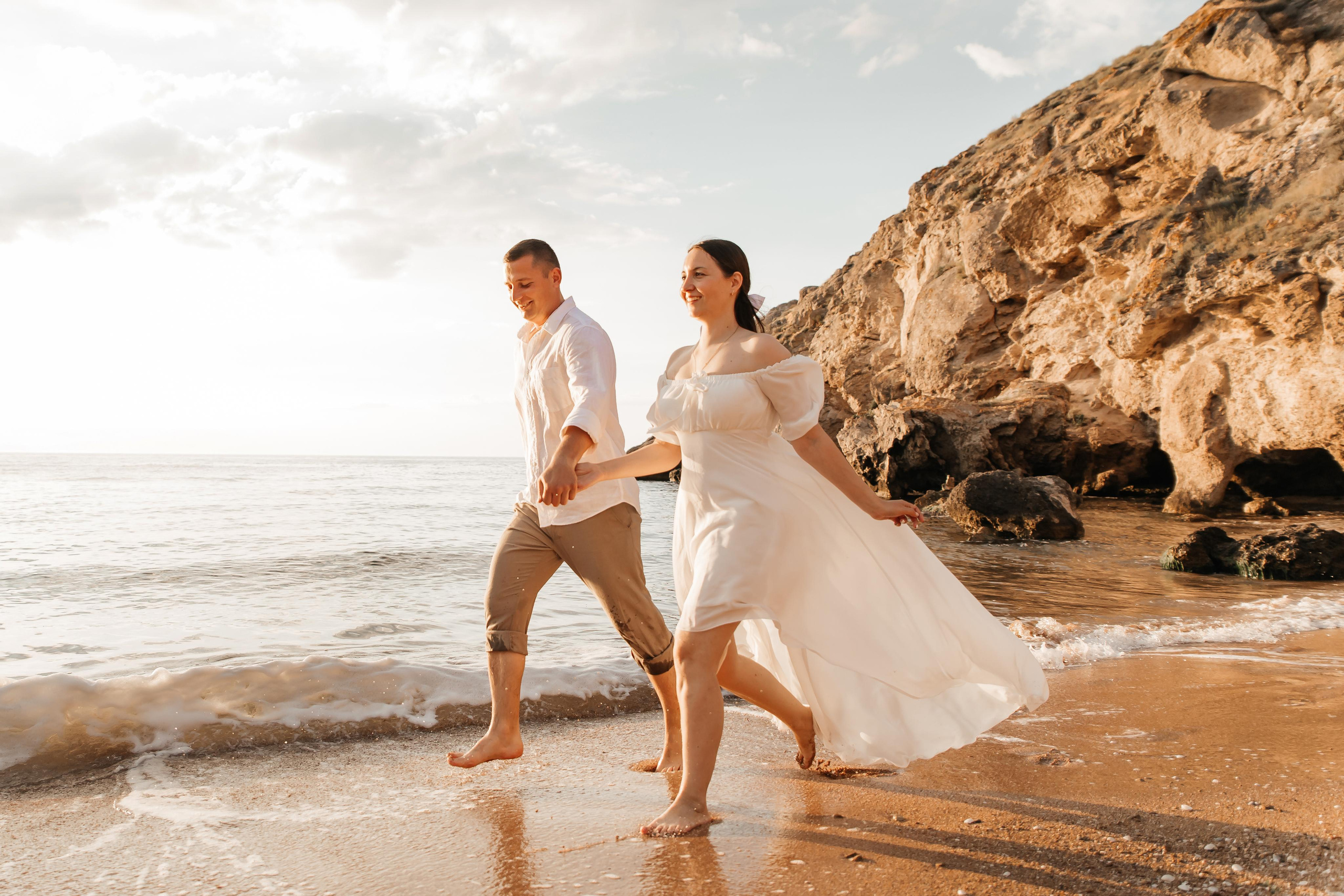 Красивая  пара крупным планом , держится за руки и бежит по морскому прибою на фоне скал. 