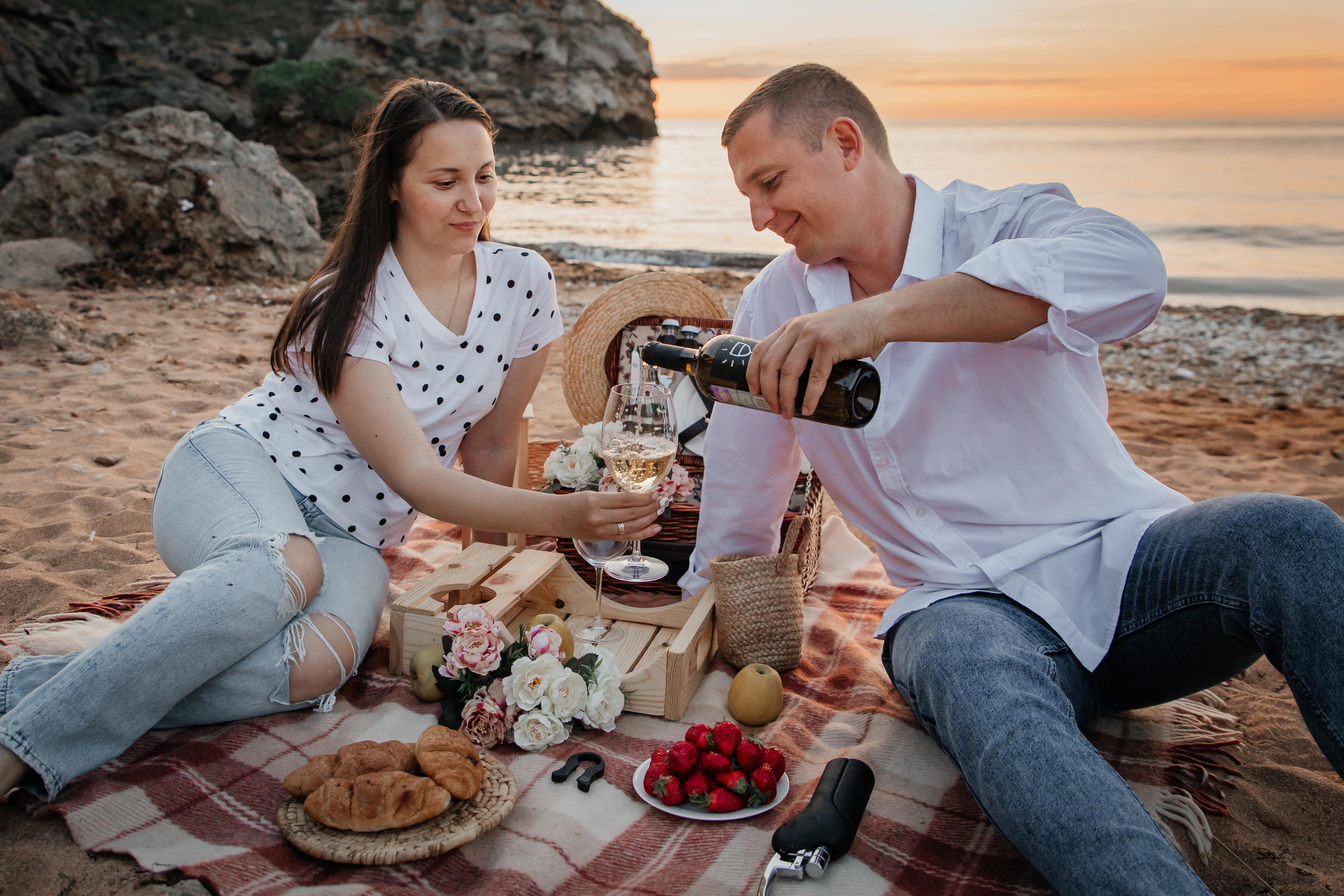 На пляже на закате парень и девушка сидят на пледе  наливают вино в бокалы . 