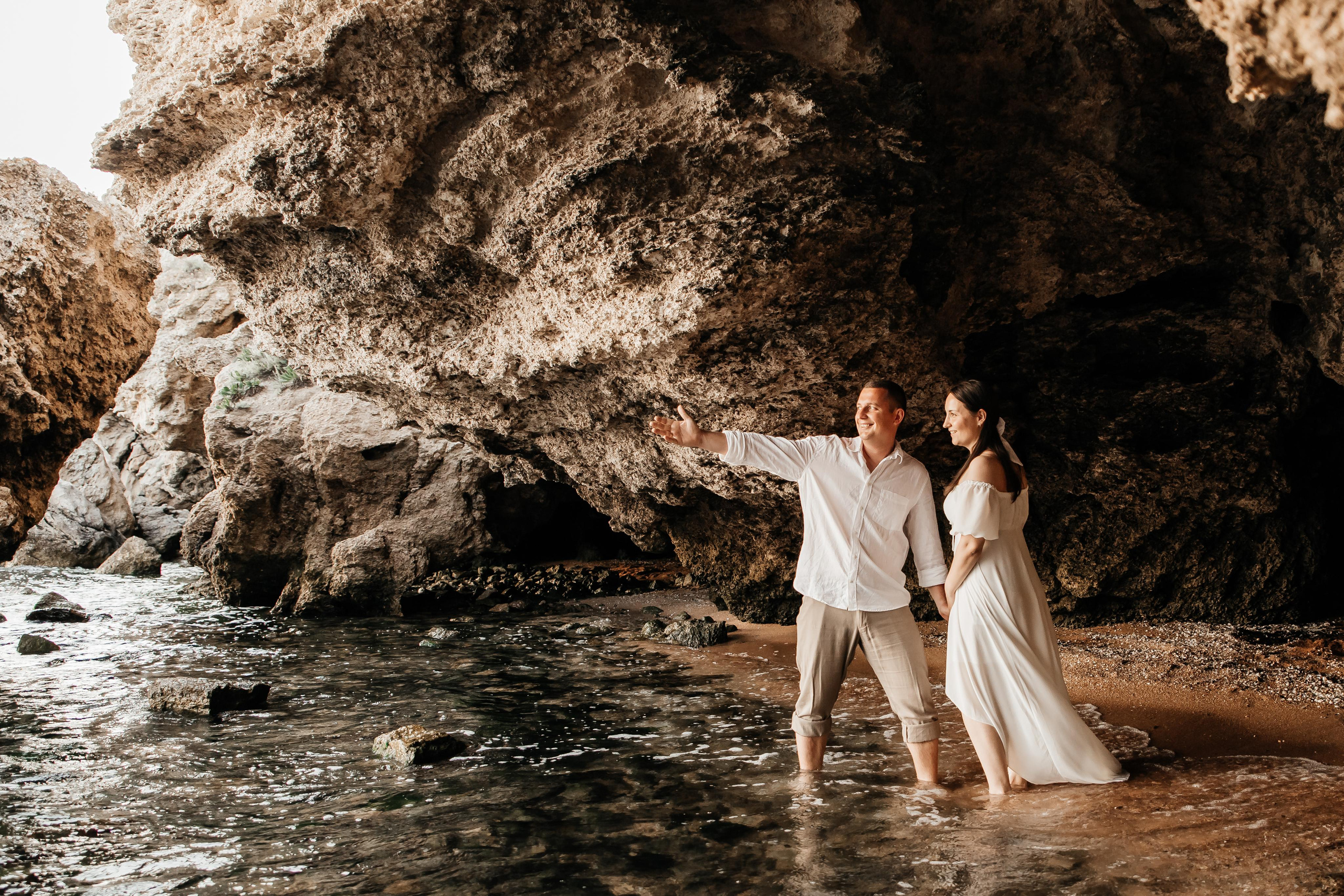 Парень показывает девушке рукой в даль, они стоят на берегу моря под скалами. 