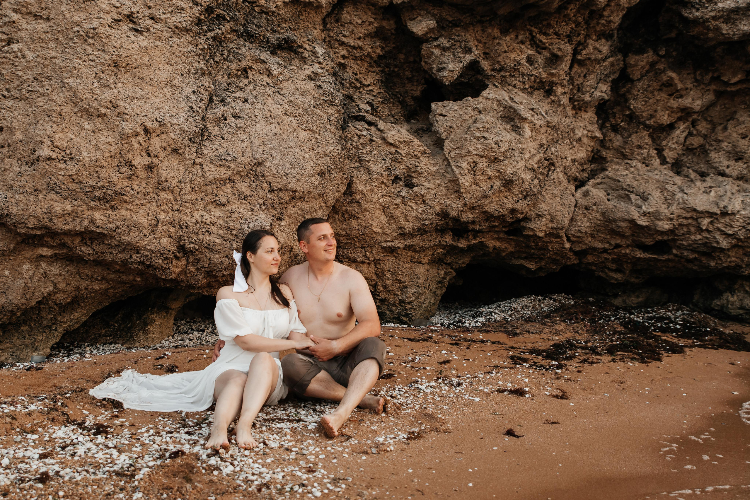 Пара в белом сидят на песке на берегу моря под скалами и смотрят вдаль. 