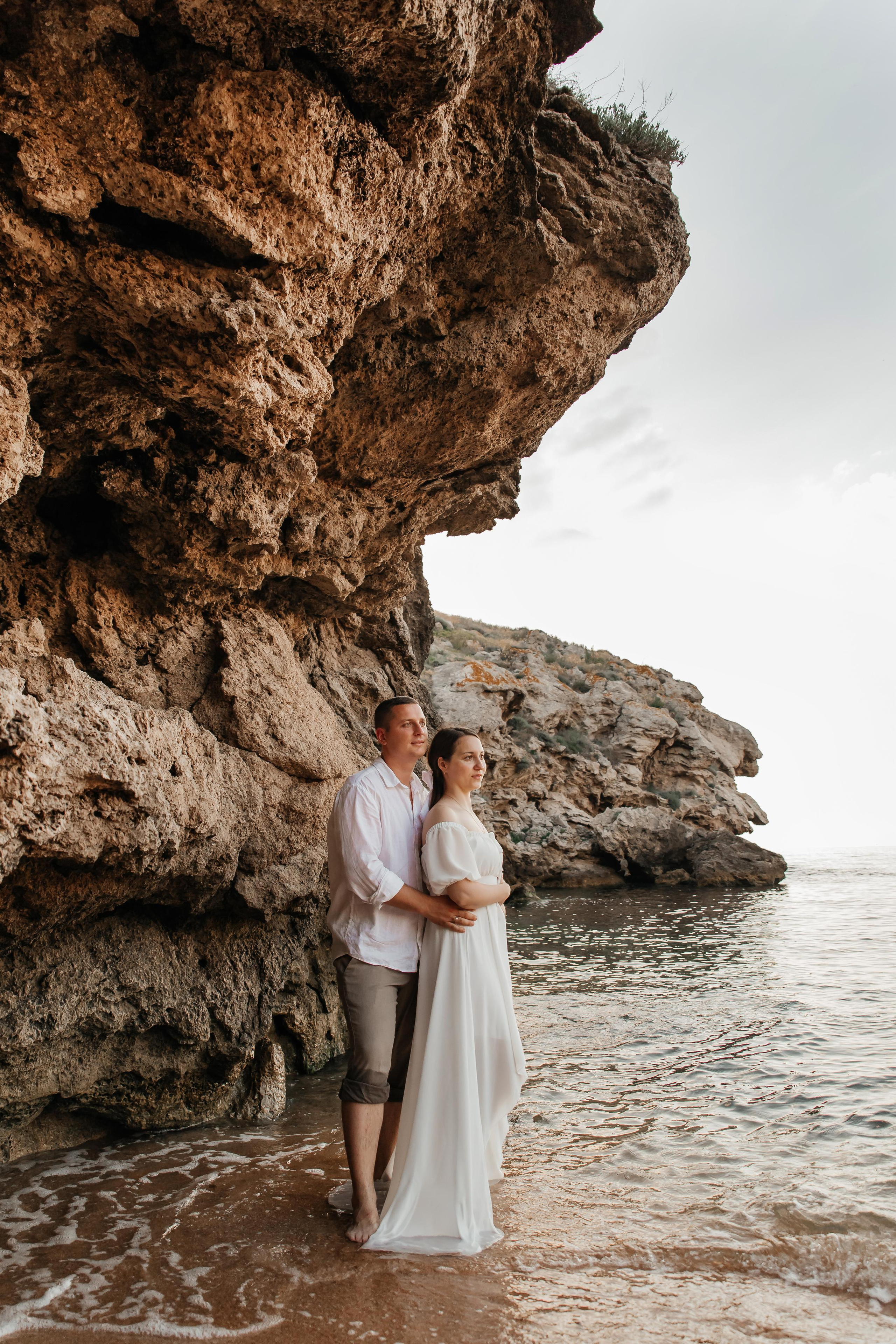 Пара в белом обнимаются и смотрят в даль, стоя  в морском прибое под скалой. 