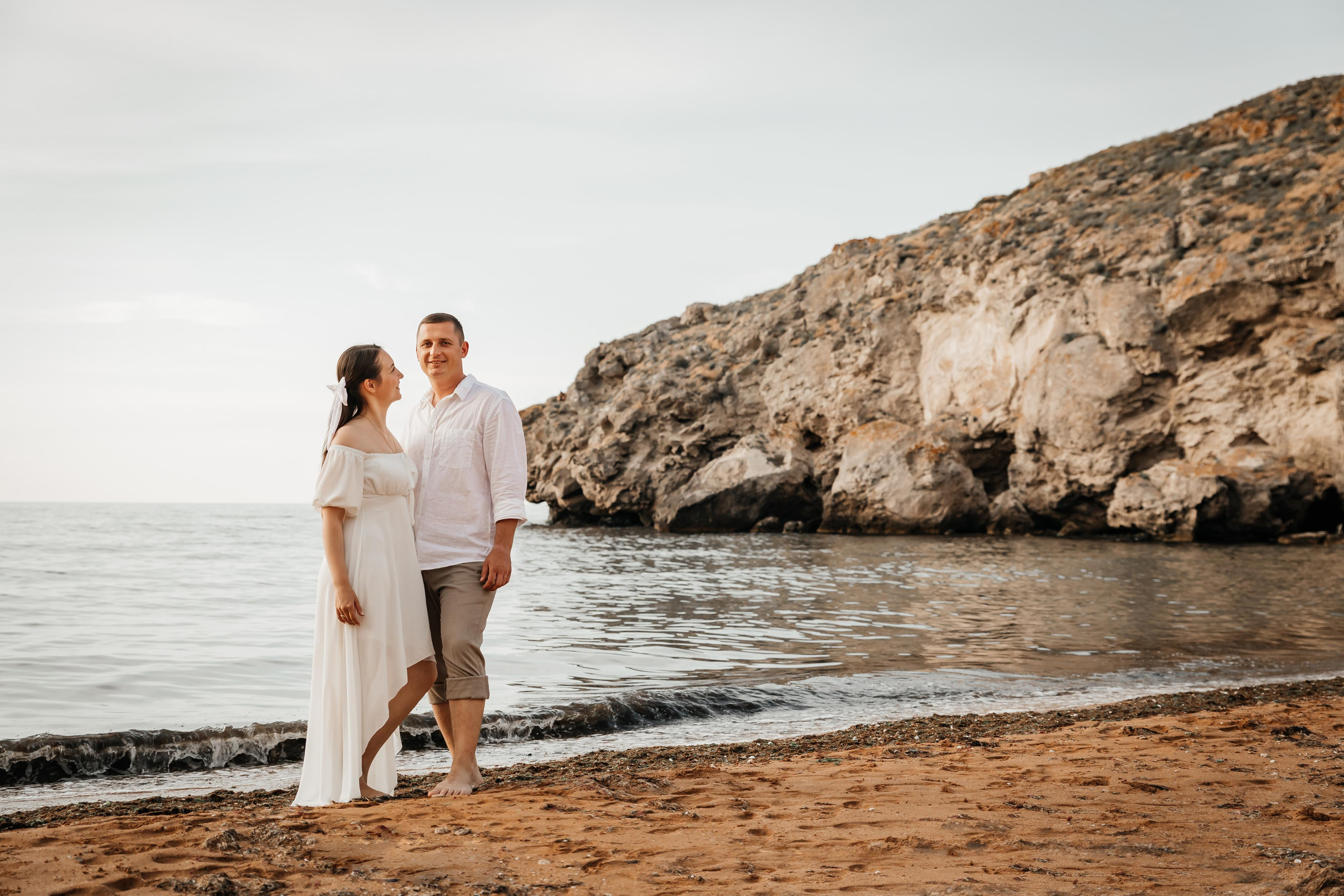 Влюбленная пара стоит на берегу моря на фоне скалы.