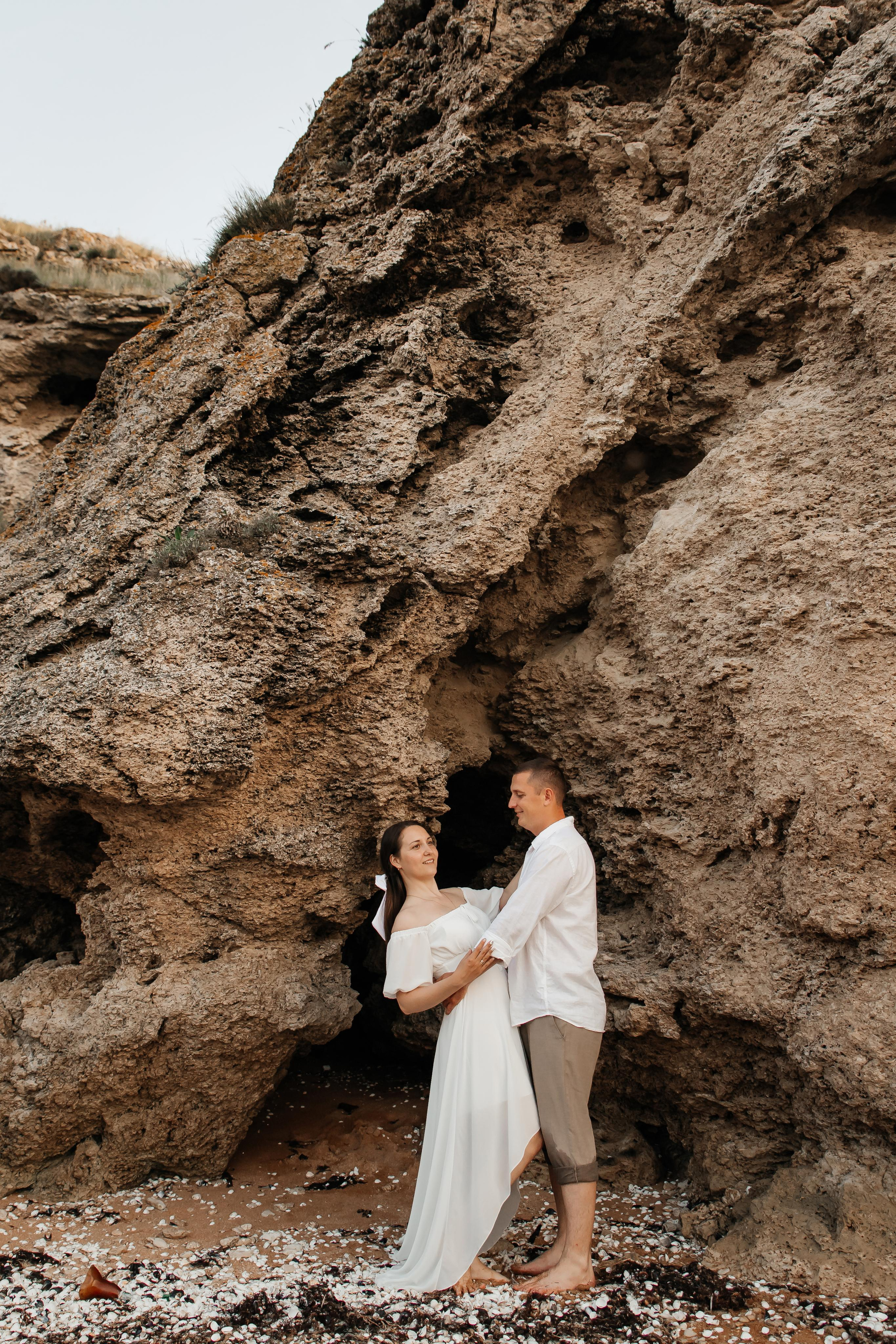 Парень и девушка в белых одеждах обнимаются на пляже под скалой. 