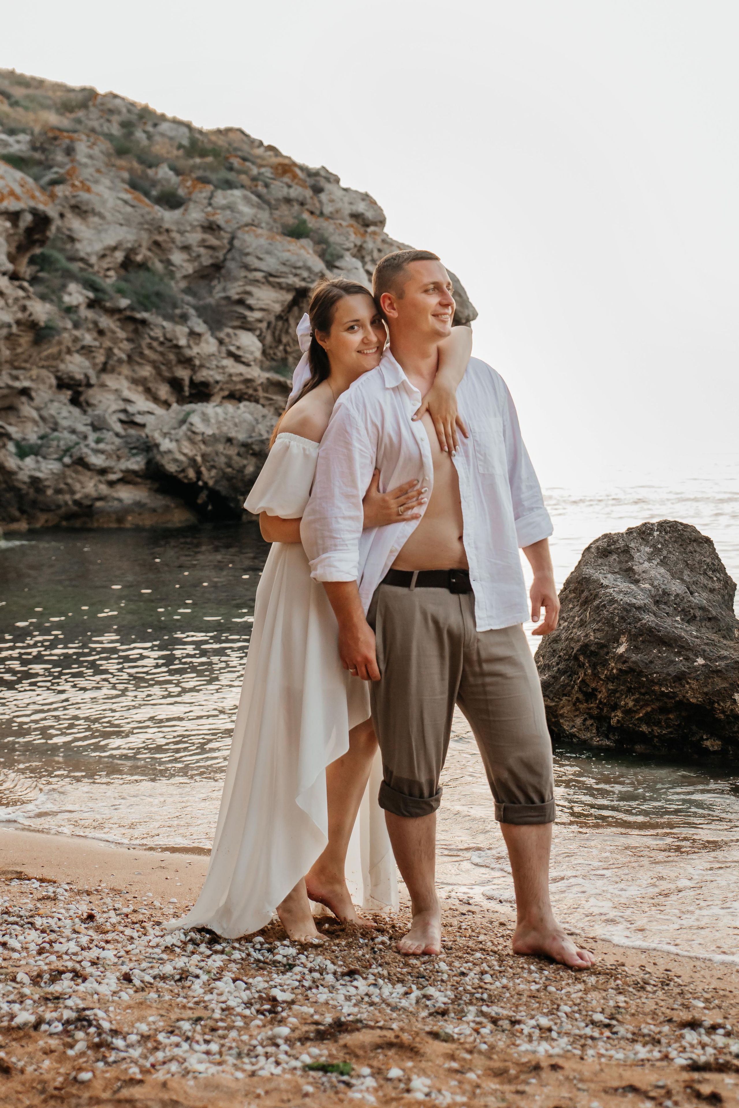 Парень смотрит в даль, сзади его обнимает девушка , они в белых одеждах на морском берегу на фоне скал. 