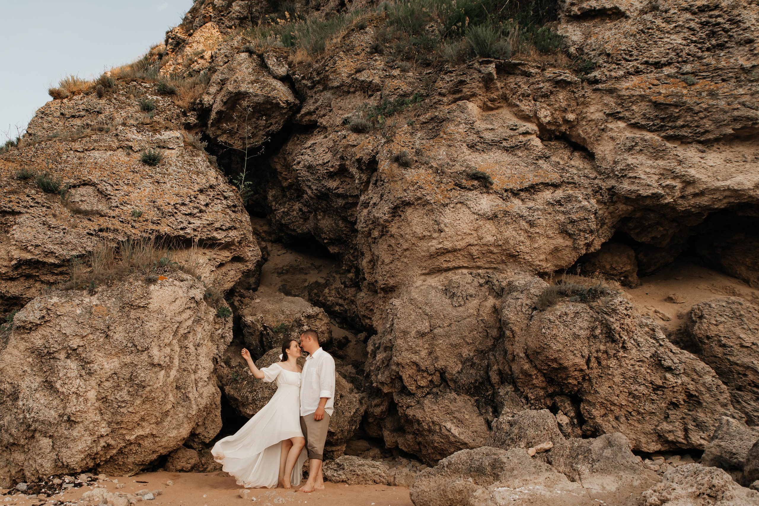 Парень и девушка в белом в летящем платье стоят на песчаном пляже под большой скалой. 
