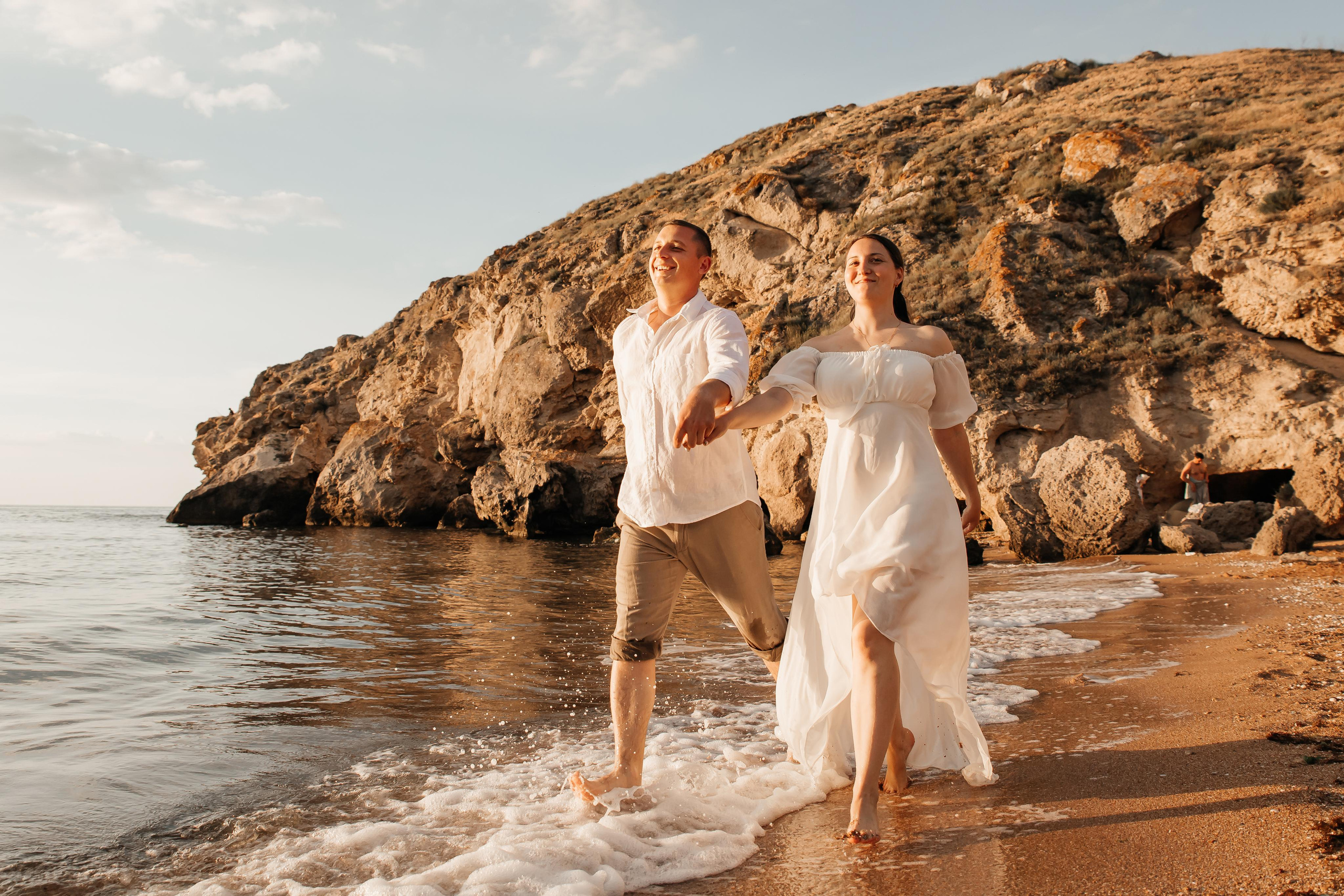 На переднем плане счастливая  пара держится за руки и бежит по морскому прибою на фоне красивых скал. 