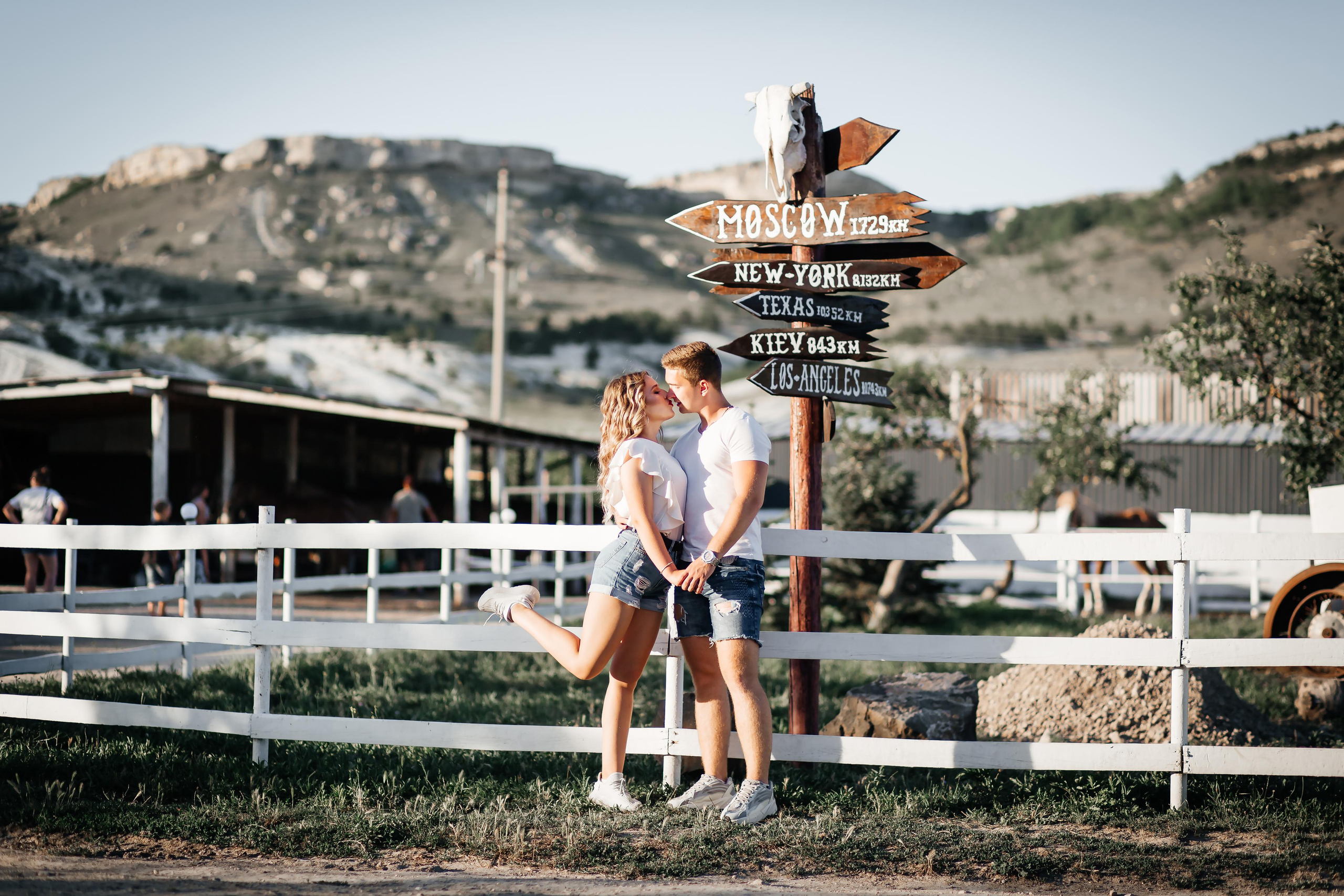 Пара целуется на фоне белого заборчика и красивого указателя дорог.
