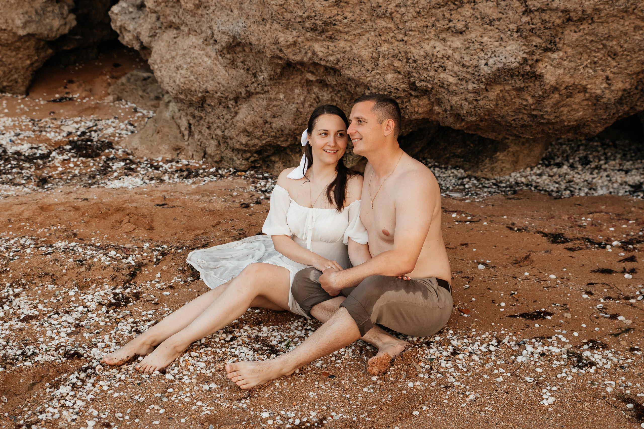 Парень и девушка сидят в обнимку на песчаном пляже под скалой. 