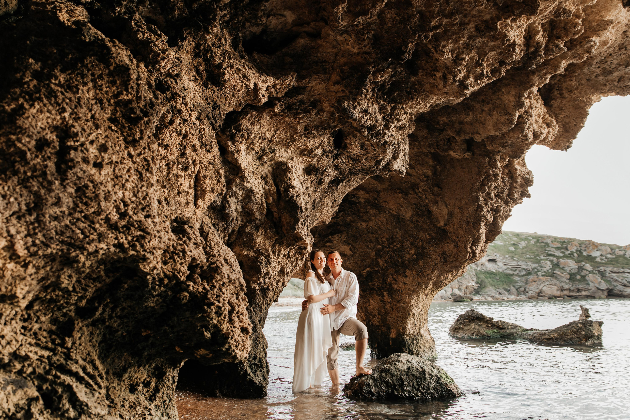 Девушка и парень стоят на берегу моря под большими скалами.