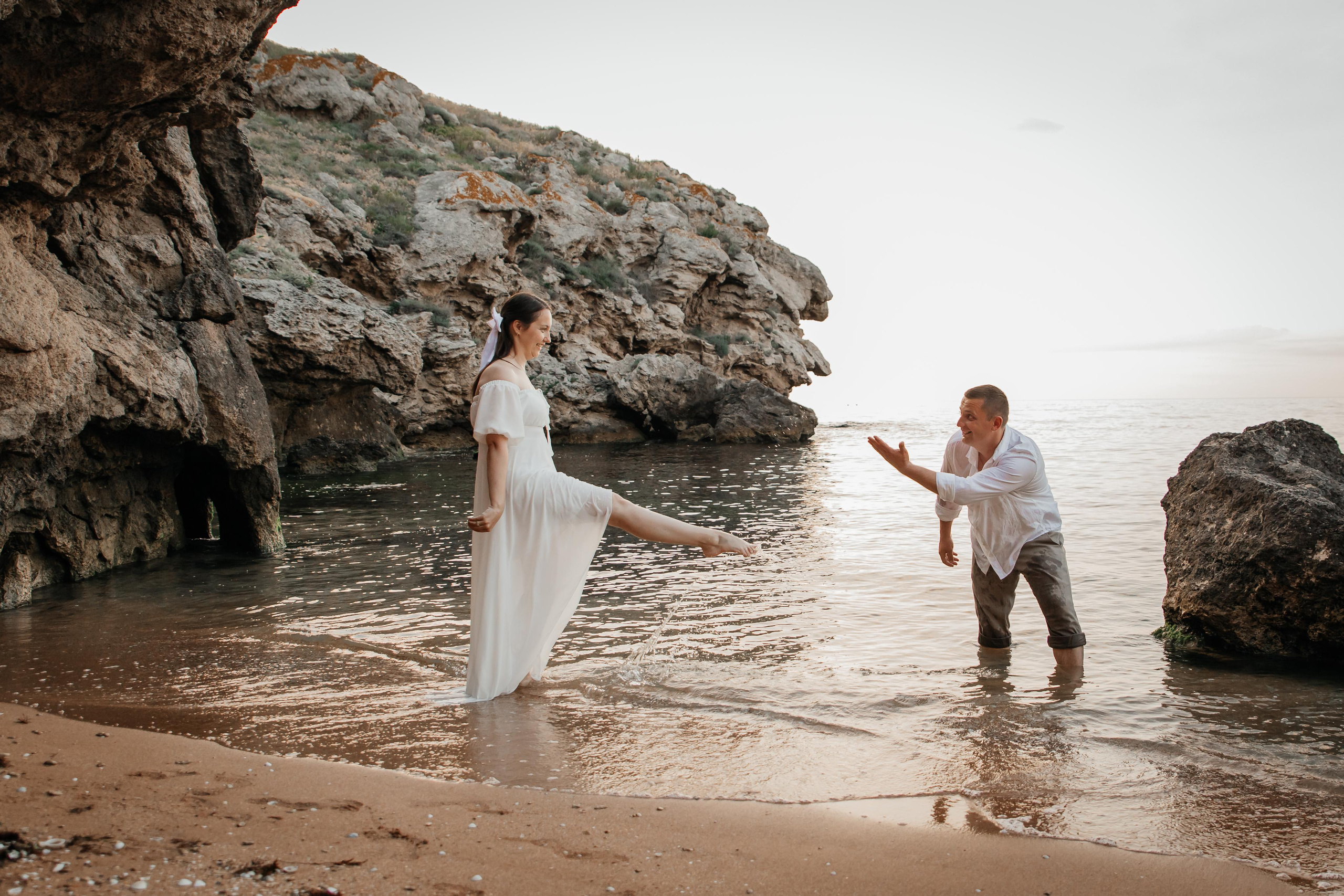 Девушка и парень на морском побережье брызгают водой друг друга на фоне скал.  
