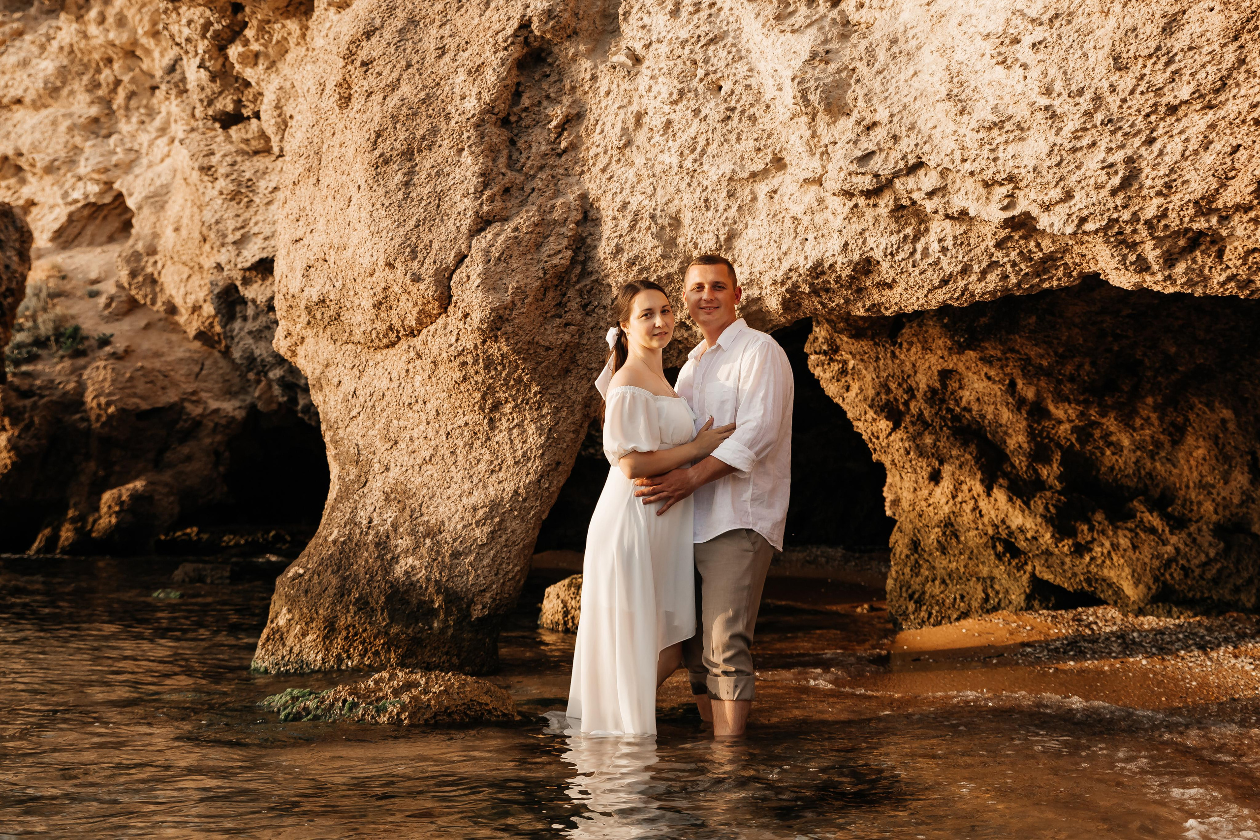 Девушка и парень в белых одеждах обнимаются стоя в море возле скал. 