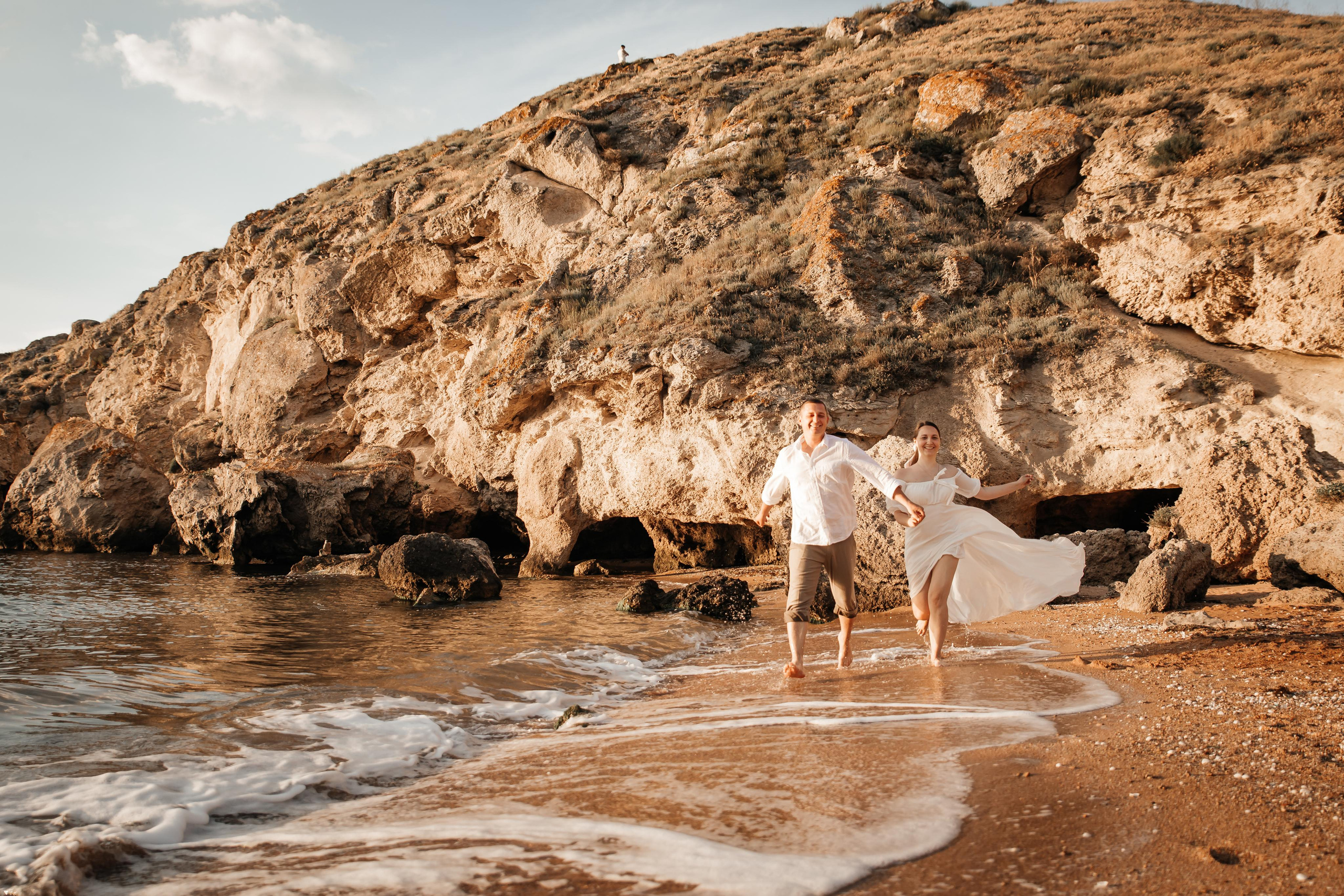 Бегущая пара в белых одеждах по морскому прибою на фоне скал. 