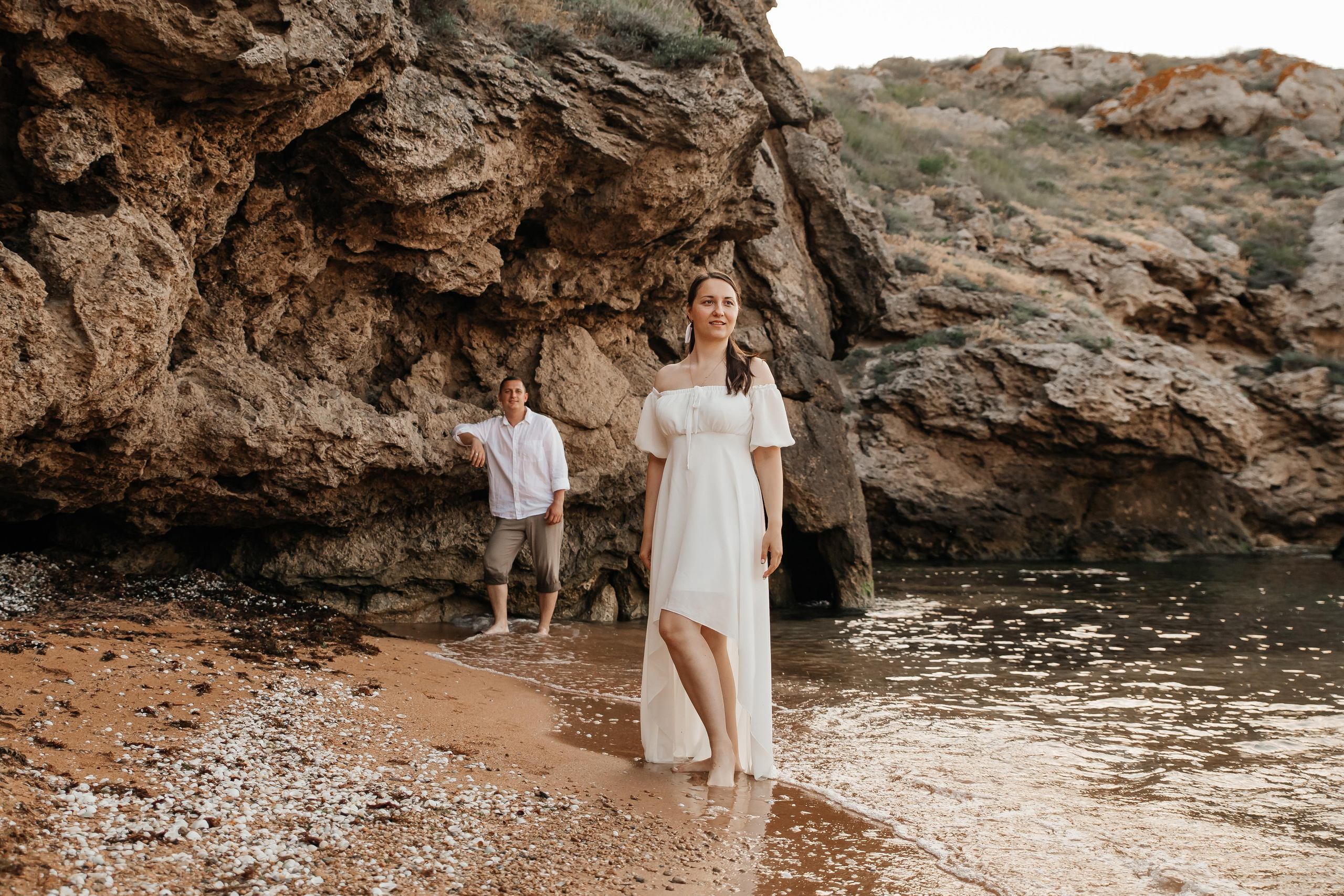 На переднем плане стоит девушка в белом платье и смотрит в даль, вдалеке стоит парень под скалой на морском берегу. 