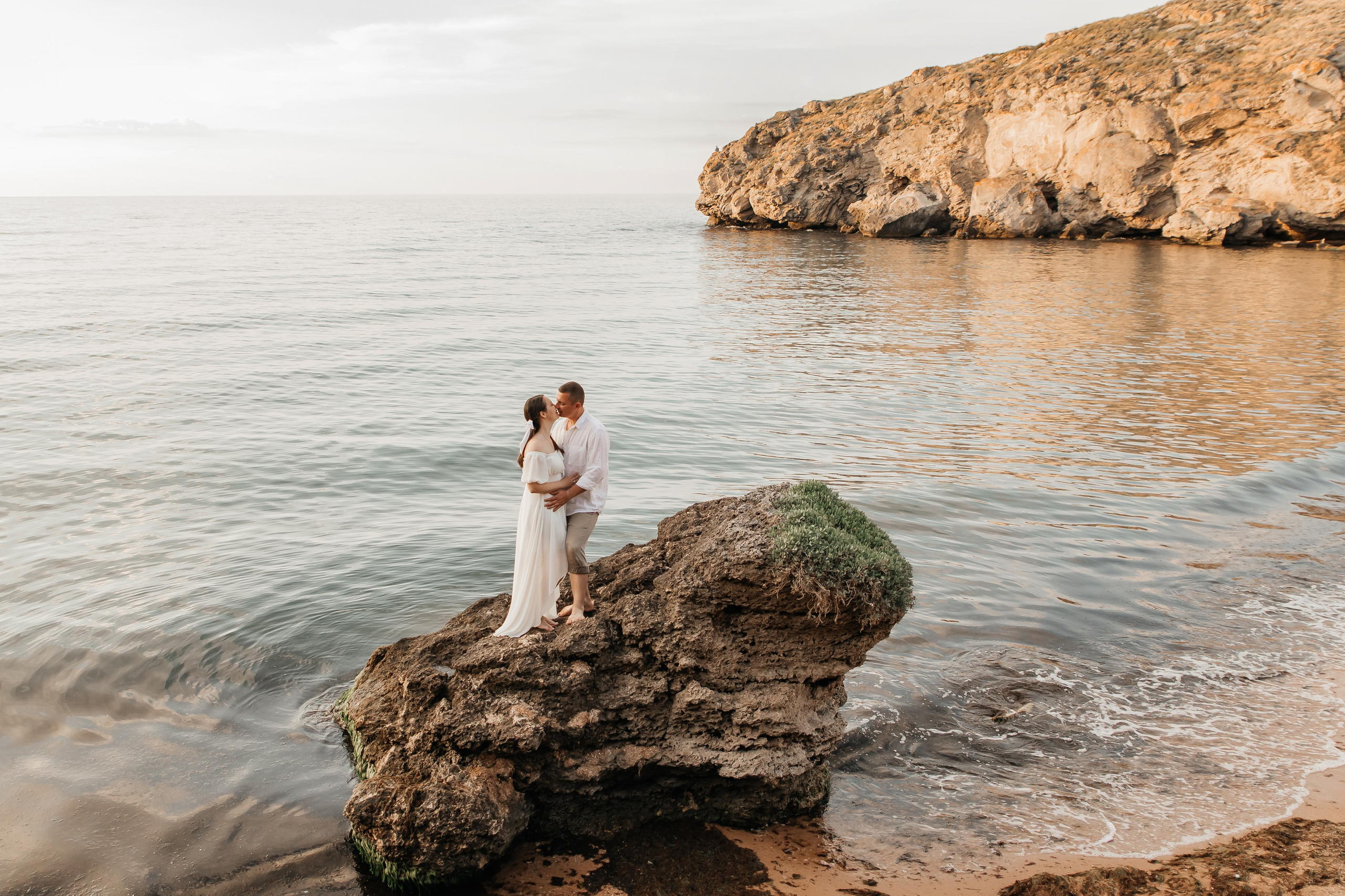 Целующаяся пара в белом на большом камне на морском берегу на фоне скал. 