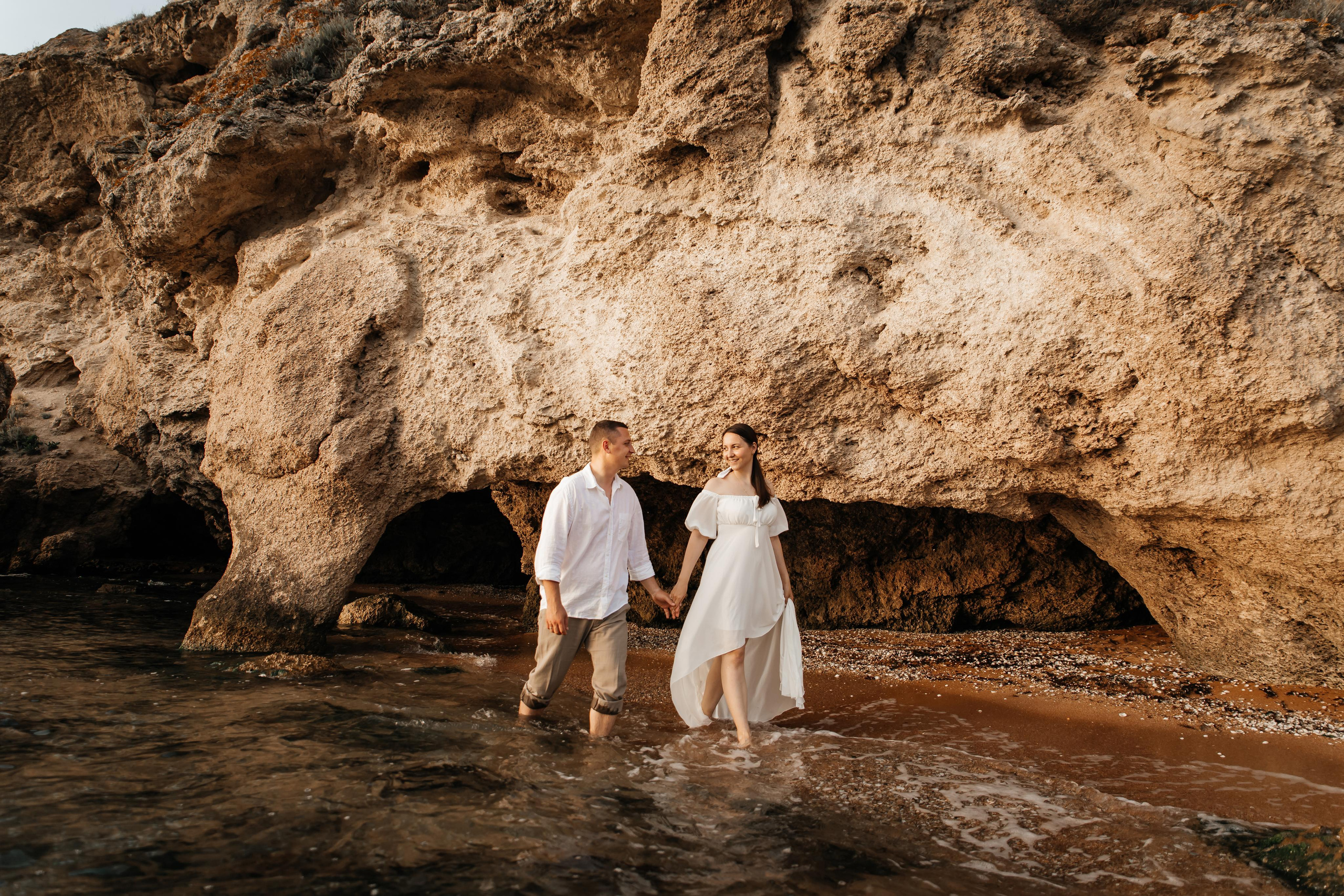 Пара идет по воде по берегу моря на фоне красивых скал. 